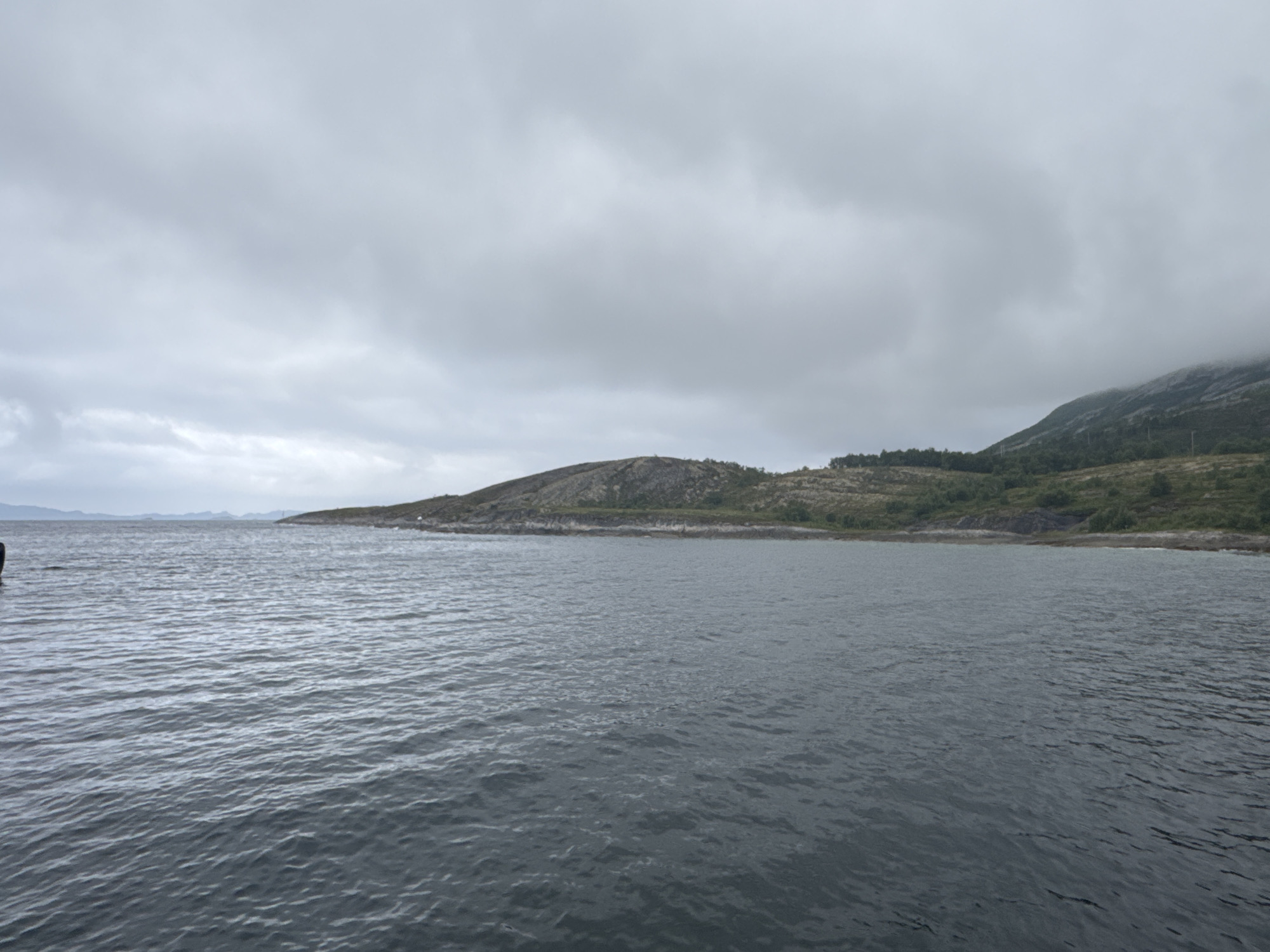 The image shows a wide, gray, choppy sea under an overcast sky. In the distance, a rocky, sparsely vegetated coastline curves into the sea. The land is covered in low-lying green shrubs and some scattered trees, with a prominent rocky outcrop dominating the center-left of the frame. Further to the right, a larger, darker hill rises, also covered in vegetation. The sky is filled with dense, gray clouds, suggesting a cool, possibly damp day. The overall mood is somber and natural, characteristic of a northern coastal environment. The scene appears to be a still photograph taken from a boat or the water's edge, looking out towards the land. There are no people or distinct objects visible in the water or on the land, emphasizing the natural landscape. The lighting is diffused and even due to the cloud cover, indicating it is likely daytime. There is no visible text in the image.