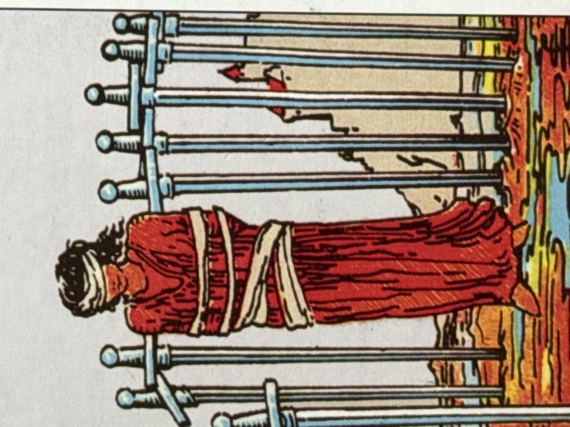 This image depicts a tarot card, specifically the Ten of Swords. The central subject is a figure lying down, seemingly asleep or incapacitated, their eyes covered with a white cloth. They are draped in a red robe that is bound with light-colored sashes. The figure is positioned horizontally, across a backdrop of ten upright swords. The swords have silver blades and light blue handles with rounded pommels. They are arranged in two rows, five above and five below the figure, creating a sense of enclosure or entrapment.
The scene is stylized and symbolic, typical of tarot card imagery. There are no clear indications of a specific time of day or weather conditions. The background behind the swords is abstract, featuring patches of light tan, dark grey, and areas with abstract shapes and colors, possibly representing distress or a tumultuous environment. On the right side of the image, there is a vertical element with fiery orange and yellow colors, along with blues and reds, suggesting intense energy or turmoil.
There is no visible text in the image. The overall mood conveyed by the card is one of suffering, completion, or rock bottom, with the figure's vulnerability emphasized by the surrounding swords. The image is presented as a still, with the orientation suggesting it might have been photographed in a vertical position.