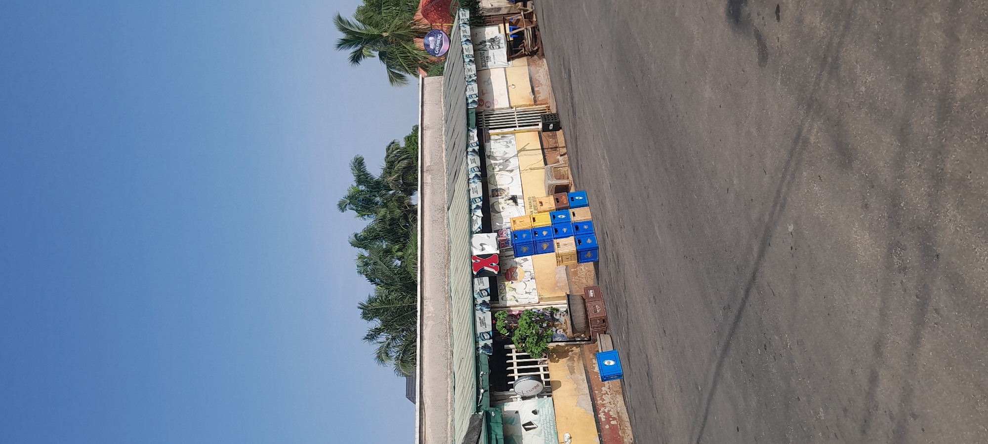A view of an outdoor establishment with a covered seating area and a collection of blue and yellow plastic crates stacked in front of it, under a bright, clear blue sky. The establishment appears to be a bar or restaurant with posters on the walls. There are palm trees visible in the background. The ground is paved, and there are various objects scattered around, including a table with some seating.