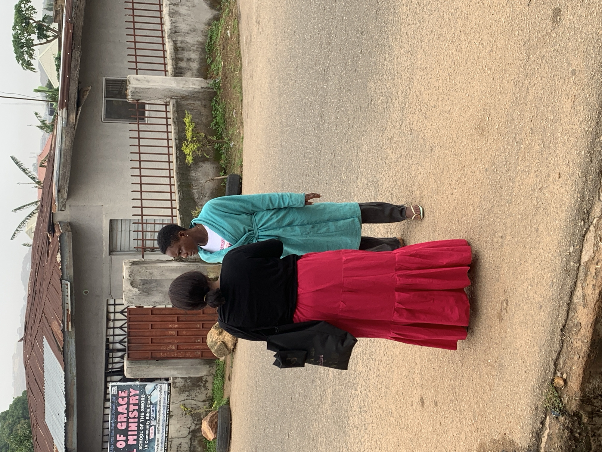 In a street scene in Jos, Nigeria, two individuals are interacting outdoors. The foreground features an asphalt road, with a curb on the left side displaying some patchy vegetation and debris. In the middle ground, two people stand on the road, facing each other. One person, from the back, wears a black long-sleeved shirt and a vibrant pink ruffled skirt. They are carrying a black tote bag over their left shoulder. The other person, facing the viewer partially, is clad in a teal blue robe over a white t-shirt and dark trousers, with one foot visible wearing a flip-flop. Behind them, a light-colored building with a weathered, corrugated metal roof and a fence made of vertical bars stands. A sign, partially visible, reads "OF GRACE SCHOOL OF THE SWORD MINISTRY". The background shows more buildings and vegetation under an overcast sky. The overall impression is of a casual, everyday street encounter in a Nigerian urban environment.