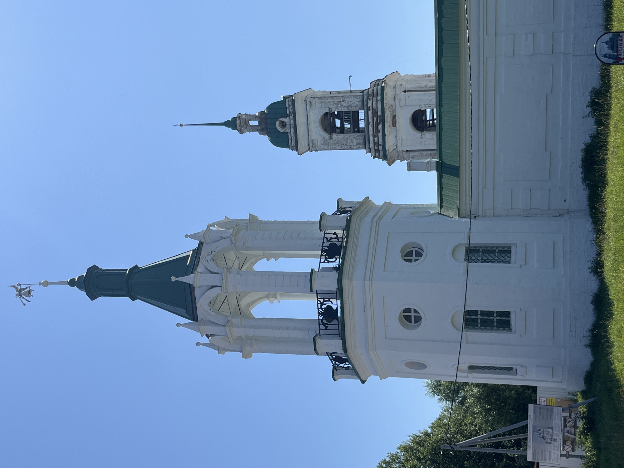 The image depicts two church bell towers against a clear blue sky. The tower in the foreground is predominantly white with a green conical roof topped by a weather vane with a figure of an angel blowing a horn. This tower features a circular base with arched windows and a balcony with black decorative railings. The second bell tower, taller and slightly to the right, is also white with a green spire and a bell visible through its open arches. This tower shows signs of wear and tear, with some weathered brickwork. In the lower part of the image, a white building with a green roof connects to the foreground tower. A grassy area with some sparse bushes is visible in front of the buildings. To the left, trees and a utility pole with a sign are present. A second sign is partially visible in the lower right corner. The scene appears to be an outdoor view of religious architecture.