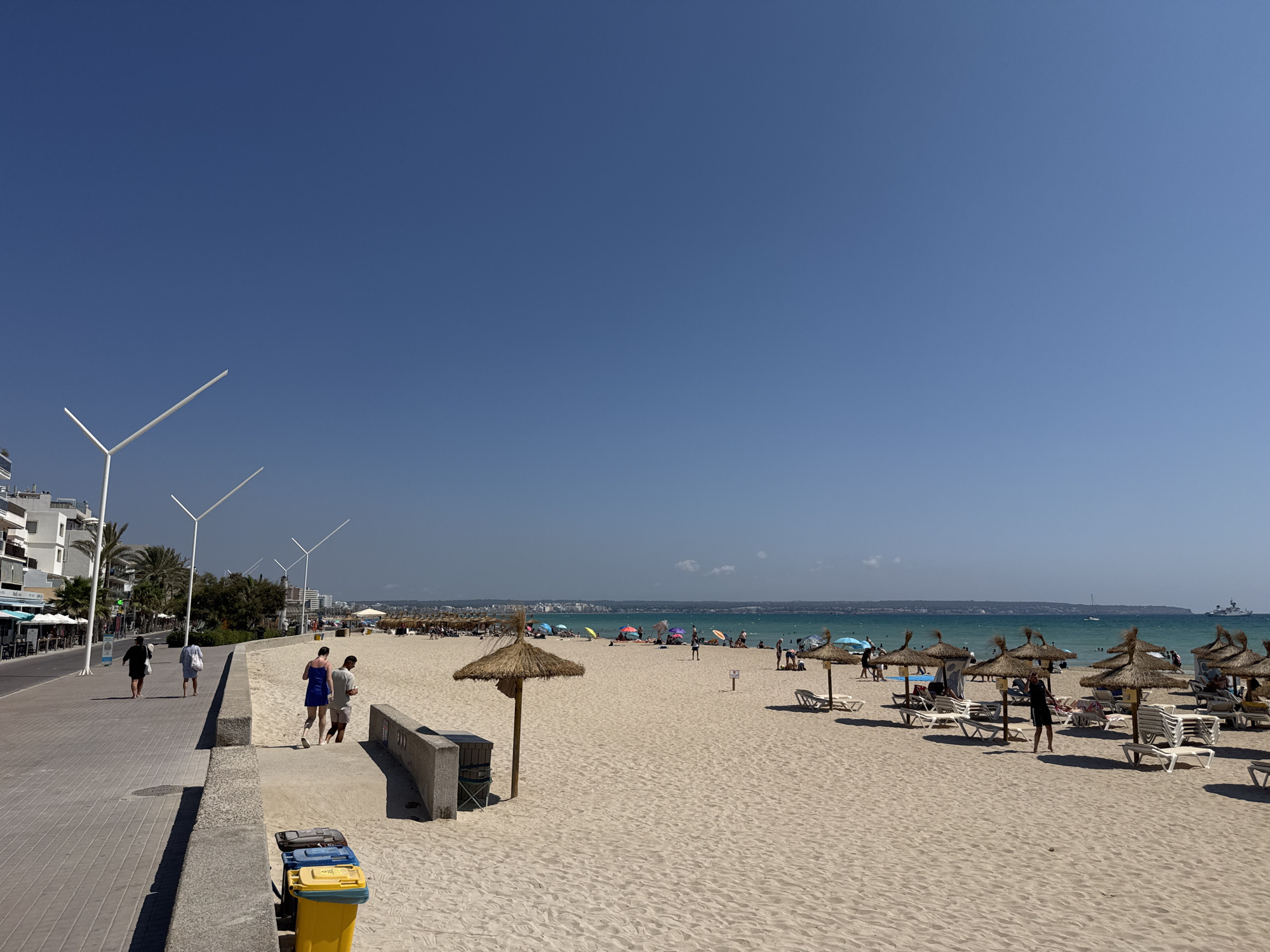 This is a daytime, eye-level, wide-angle shot of a beach promenade and the beach area in Palma de Mallorca, Spain. The scene is brightly lit under a clear blue sky. On the left, a paved promenade runs along the edge of the beach. Several people are walking on the promenade, some in casual attire. Buildings with balconies and awnings line the promenade. A few palm trees are visible.
The beach itself is covered in light-colored sand and is dotted with numerous straw umbrellas and white lounge chairs, many of which are occupied by beachgoers. The turquoise water of the sea stretches to the horizon, where a hazy coastline and a few distant boats are visible. Some individuals are seen walking on the beach and in the shallow water. A small concrete wall separates the promenade from the beach in some sections. Two sets of trash bins are located on the promenade near the edge of the beach. The overall atmosphere suggests a sunny, active day at a popular tourist destination.