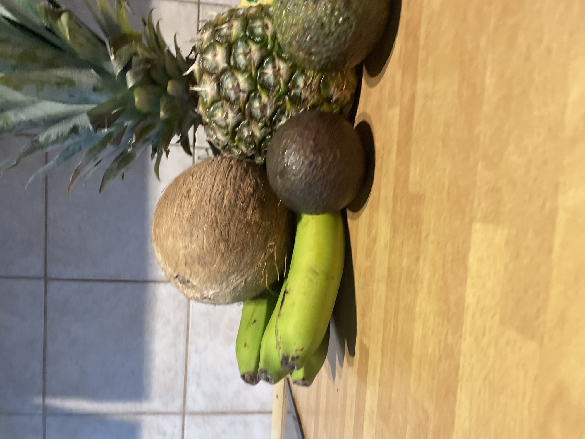 The image shows a variety of fruits arranged on a wooden countertop in front of a tiled wall. The fruits include a whole coconut, a pineapple, a bunch of green bananas, and two avocados. The coconut is positioned behind the bananas and to the left of the pineapple. The pineapple is located to the right of the coconut. Two avocados are visible in the foreground, one to the left and slightly in front of the pineapple, and the other to the right and slightly behind the first avocado. The wooden countertop has a natural grain pattern. The tiled wall in the background appears to be light-colored with dark grout lines. The lighting suggests an indoor setting. No people or actions are depicted.