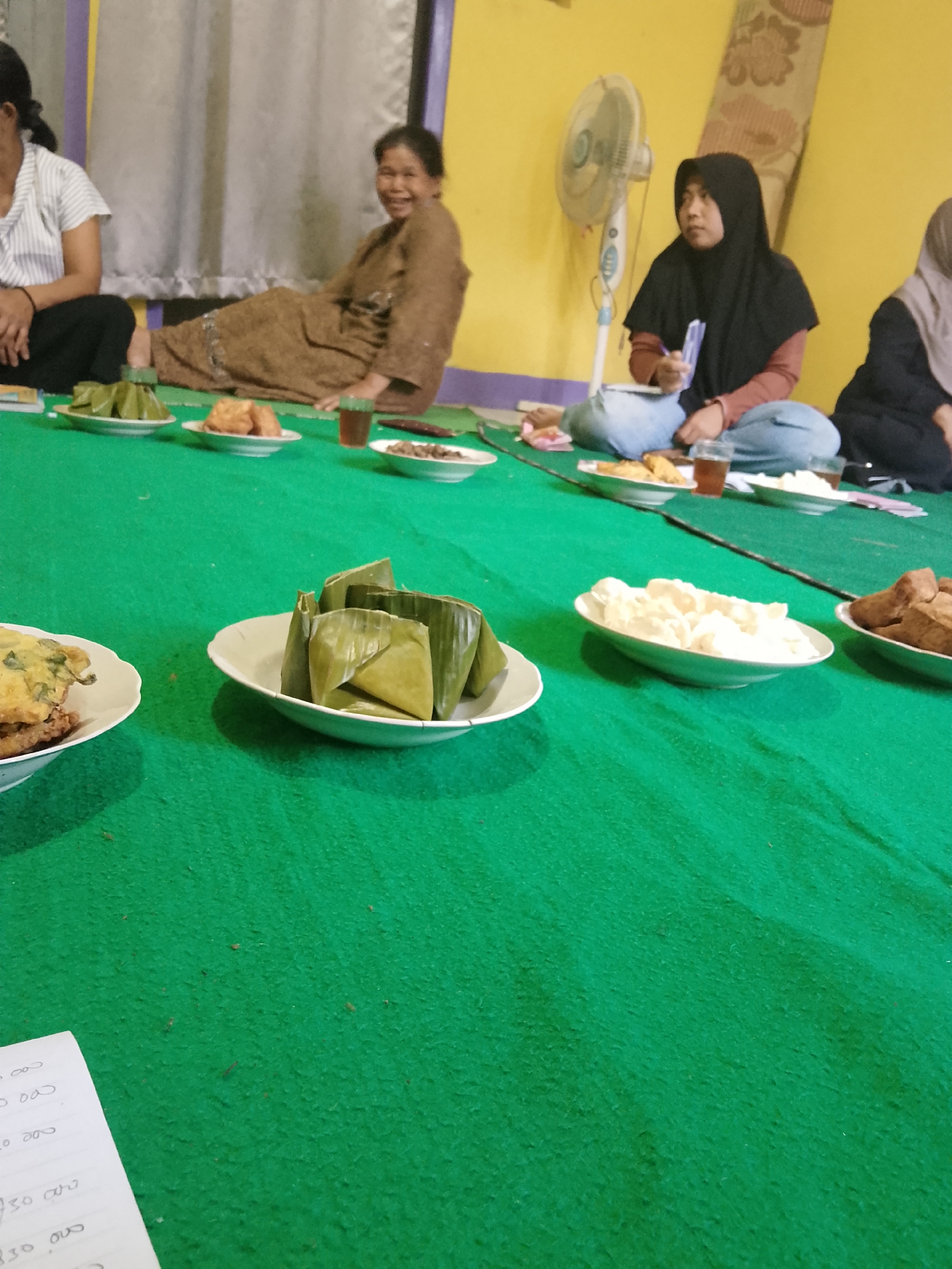 In a room with yellow walls, several individuals are seated on a green carpet. A woman on the left is wearing a white striped shirt and dark pants, her left arm resting on her lap. To her right, a woman in a brown dress is seated, smiling and looking forward. Further to the right, a person wearing a black hijab and a reddish-brown top is seated, holding a piece of paper and a pen. Another person in a light-colored hijab is partially visible on the far right.
Arranged on the green carpet in front of the seated individuals are several plates of food. One plate contains several items wrapped in green leaves. Another plate holds fried items, and a third contains white, possibly fluffy food. A glass of dark liquid is also present on the carpet. A standing fan is visible in the background. A piece of paper with handwritten numbers, possibly a list or receipt, is in the lower left corner of the image, resting on the green carpet. The scene appears to be indoors, in Jumo, Indonesia.