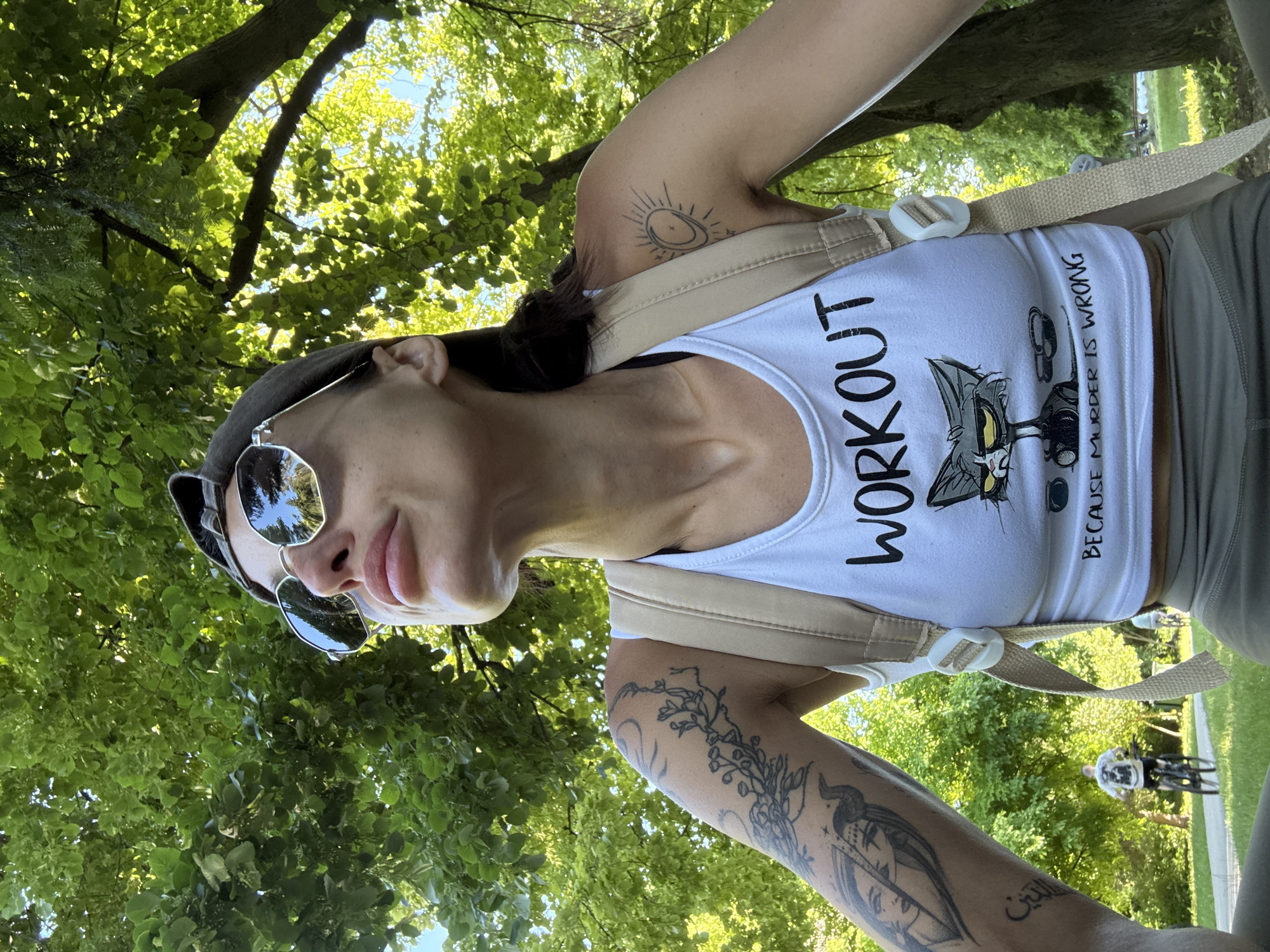 A woman with tattoos on her arms is wearing sunglasses, a baseball cap, and a white tank top with the words "WORKOUT" and a cartoon cat graphic. She is outdoors, with trees and greenery surrounding her. In the background, two individuals are seen cycling on a path. The location is the Kaylaka Park in Pleven, Bulgaria.