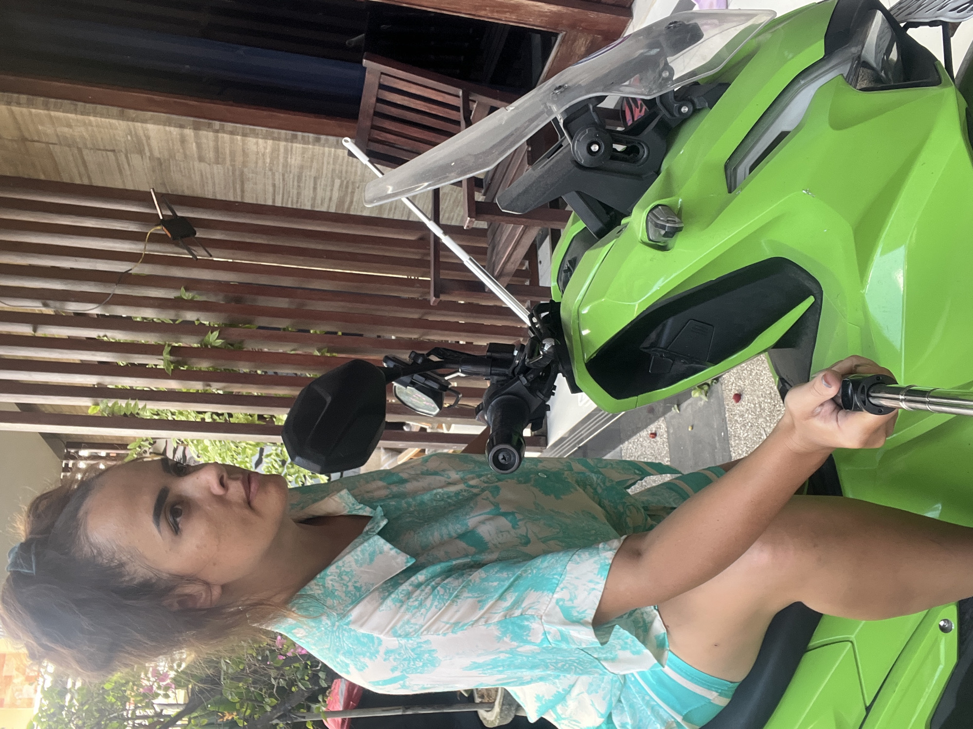 A woman is sitting on a bright green scooter in Kuta Selatan, Indonesia. She is wearing a light blue and white patterned short-sleeved collared shirt and matching shorts. Her dark hair is pulled up in a messy bun with a blue scrunchie. She is looking to her left, away from the camera, and is holding a selfie stick with her right hand. The scooter has a clear windshield and black handlebars. In the background, there is a wooden fence and a brown wooden chair. Green foliage and pink flowers are visible to the left of the woman. The ground is paved with stone tiles.