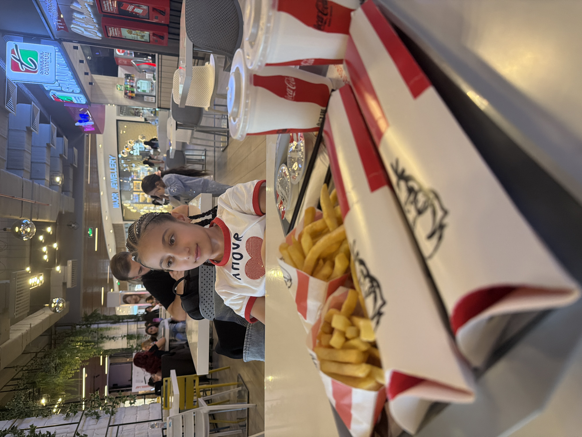 The photograph captures a young girl seated at a table in what appears to be a food court or restaurant, possibly in Yerevan, Armenia, as suggested by the presence of a sign with Armenian script in the background. In front of the girl, on a tray, are two large servings of french fries in containers with the KFC logo, accompanied by at least two Coca-Cola beverages in plastic cups with lids and straws.
The girl is wearing a white shirt with the word "amour" and a heart design. An adult male is seated beside her. The surrounding area is populated with other patrons at tables and chairs, indicating a bustling environment. The decor includes modern lighting fixtures and green plants along a wall, suggesting an indoor location, likely a shopping mall. The lighting is bright and artificial, casting a slight glare on the tray's surface.
