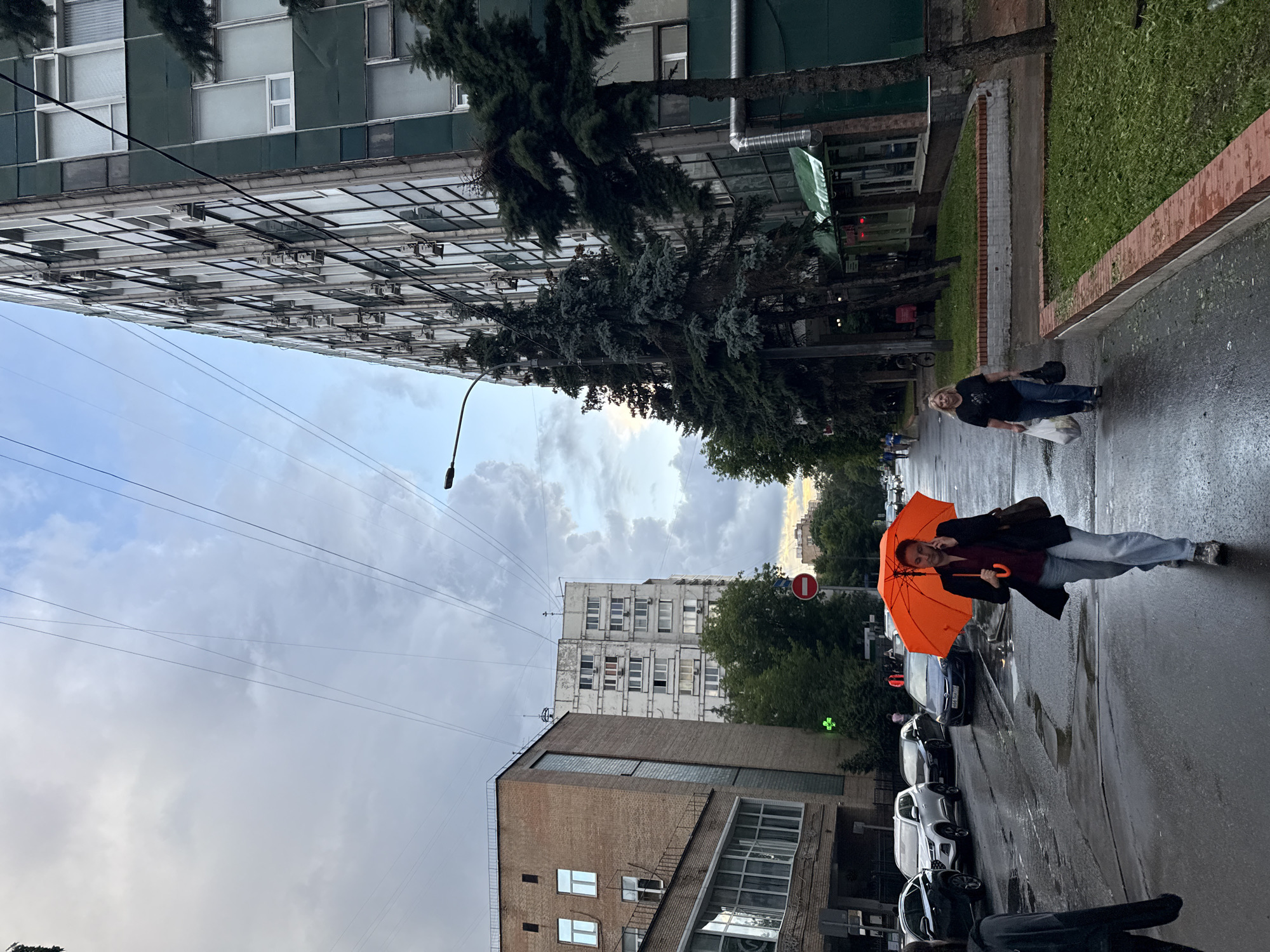 An overcast sky looms over a wide, wet street in Moscow, Russia. A woman walks towards the camera, holding an open orange umbrella and speaking on her phone. She is wearing a dark jacket over a burgundy shirt and light-colored jeans. To her right, another woman walks in the same direction, carrying two plastic bags. She is wearing a black t-shirt and blue jeans. A line of parked cars is visible on the left side of the street, and tall, multi-story buildings line the right side. One building on the right is particularly prominent, with a dark green facade and numerous windows. Coniferous trees line the sidewalk in front of this building. The street surface is wet and reflective, indicating recent rain.