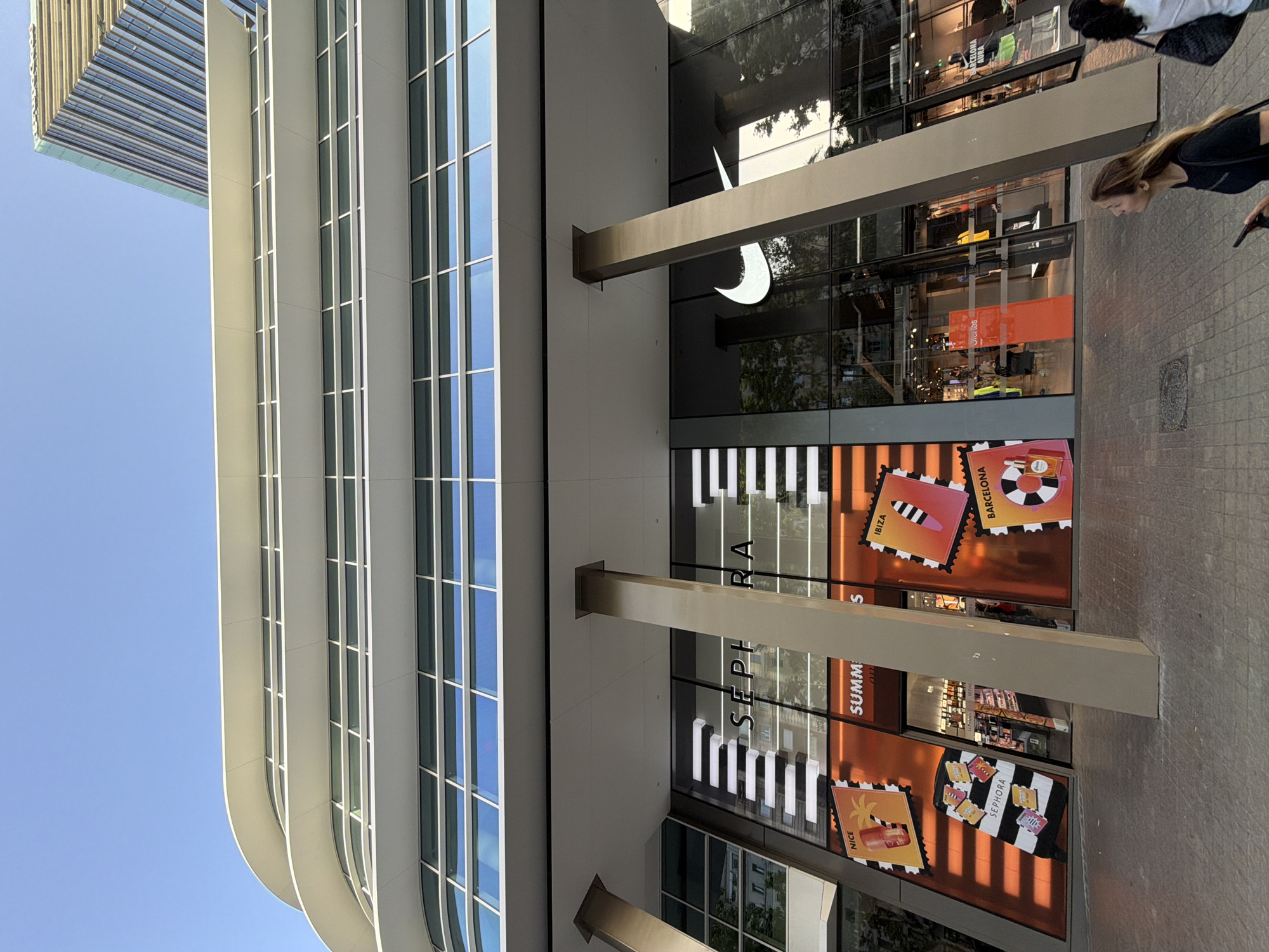 This is a low-angle, outdoor, daytime photograph of the facade of a multi-story modern building in Barcelona, Spain. The building features a white, curved upper structure with multiple horizontal bands of windows. Below this, the ground floor is predominantly glass, housing two retail stores. The store on the left is Sephora, identifiable by its prominent signage and decorative window displays featuring images reminiscent of travel stamps with destinations like "Nice," "Ibiza," and "Barcelona." One display depicts a suitcase adorned with "Sephora" stickers. The store to the right is a Nike store, indicated by a large, illuminated Nike swoosh logo on its glass facade. The entrance to the Nike store is visible, with mannequins and merchandise displayed inside. The ground surface is paved with large, rectangular tiles. In the foreground, two women are walking on the sidewalk. The woman on the right is looking upwards and to her left, while the woman on the left is partially visible, holding a phone. The sky is clear and blue.