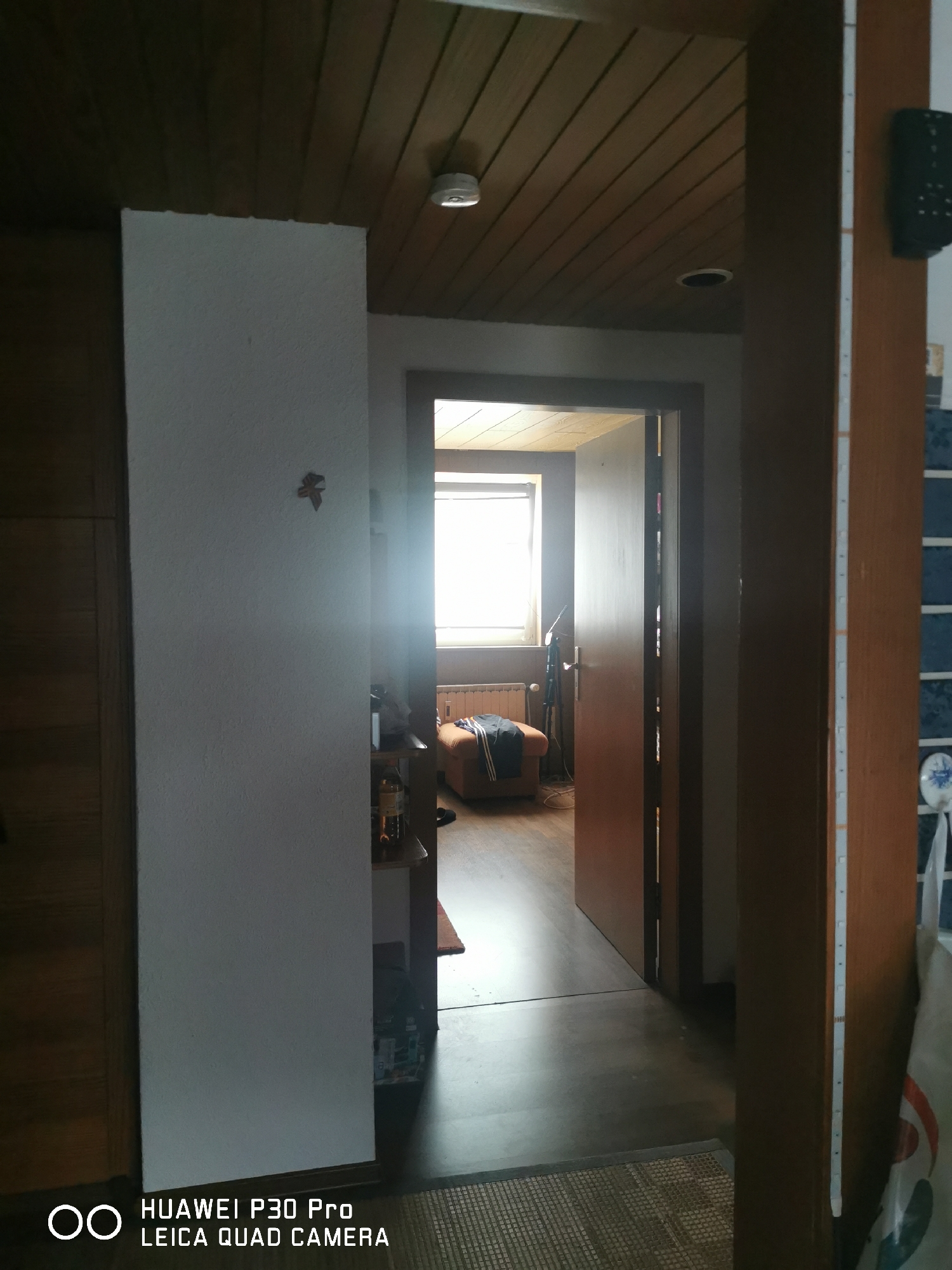 The image shows an interior view of a hallway and an open doorway leading to another room. The hallway features a white textured wall on the left, a wooden ceiling with visible planks, and a wooden door frame on the right. A small decorative item resembling a leaf is affixed to the white wall. The doorway reveals a glimpse into a room with a window, a radiator, and a brown armchair with clothing draped over it. The floor in the hallway appears to be dark wood or laminate, and a woven mat is visible in the foreground. The overall lighting is dim, with a bright light source emanating from the window in the adjacent room. The image includes a watermark at the bottom indicating "HUAWEI P30 Pro LEICA QUAD CAMERA". This image was taken on October 15, 2023, at 14:59. The metadata does not contain specific location information for Bad Oeynhausen, Germany.