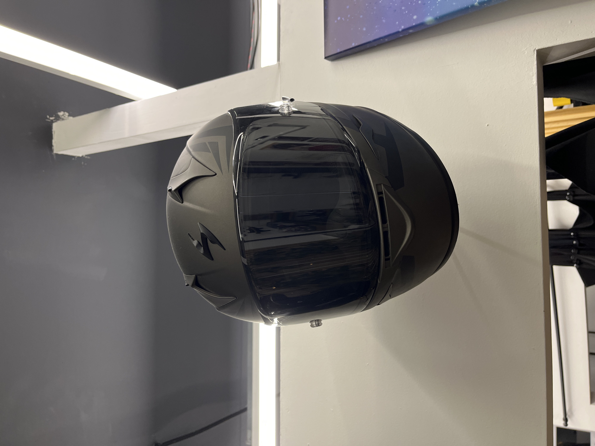 A matte black Scorpion brand full-face motorcycle helmet with a tinted visor is displayed, suspended from the ceiling by an unseen mechanism. The helmet is angled slightly to the left and tilted forward. It features the Scorpion logo, a stylized "S", in black on the top. Gray geometric graphics are visible on the sides of the helmet. The helmet is positioned against a white wall with a gray ceiling above. Two long, thin fluorescent light fixtures are mounted on the ceiling, one on the left and one on the right, illuminating the scene. A portion of a framed artwork depicting a starry sky is visible on the right side of the image. The context of the image is a retail environment, possibly a motorcycle shop. There are no people or animals present in the image. The location is Chaikovsky, Russia.