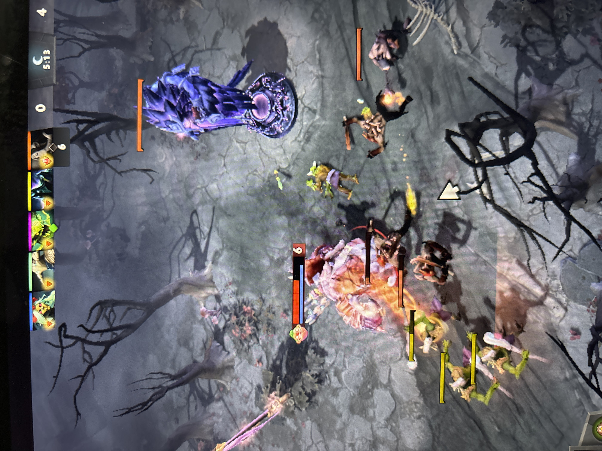 A screenshot from a fantasy-themed video game, likely Dota 2, depicts a combat scene within a dark, overgrown environment, viewed from a top-down, isometric perspective.
In the upper left portion of the image, a large, purple crystalline structure, resembling a tower, stands with an orange health bar above it. To its right, several smaller, humanoid combat units, appearing to be "creeps" from two opposing factions, are engaged in battle. Near the bottom-center, a more prominent hero character with a large, glowing weapon is surrounded by these creeps, displaying a level '9' indicator on its health and mana bars. Another character or larger unit is visible further to the right, also involved in the skirmish.
The game's user interface is present on the left side, featuring a vertical bar that includes a moon icon, the game time "5:13", a score of "0", and a stack of hero portraits. One of these portraits shows a level '6'. Various units across the battlefield display health bars in orange, yellow, red, and blue. A white arrow cursor points towards the center-right of the screen. The environment is characterized by dark, twisted trees and skeletal remains, contributing to a somewhat gloomy atmosphere. The "5:13" time and moon icon suggest an in-game night cycle. The image itself appears to be a photograph of a display screen.
Location context: Sofia, Bulgaria.