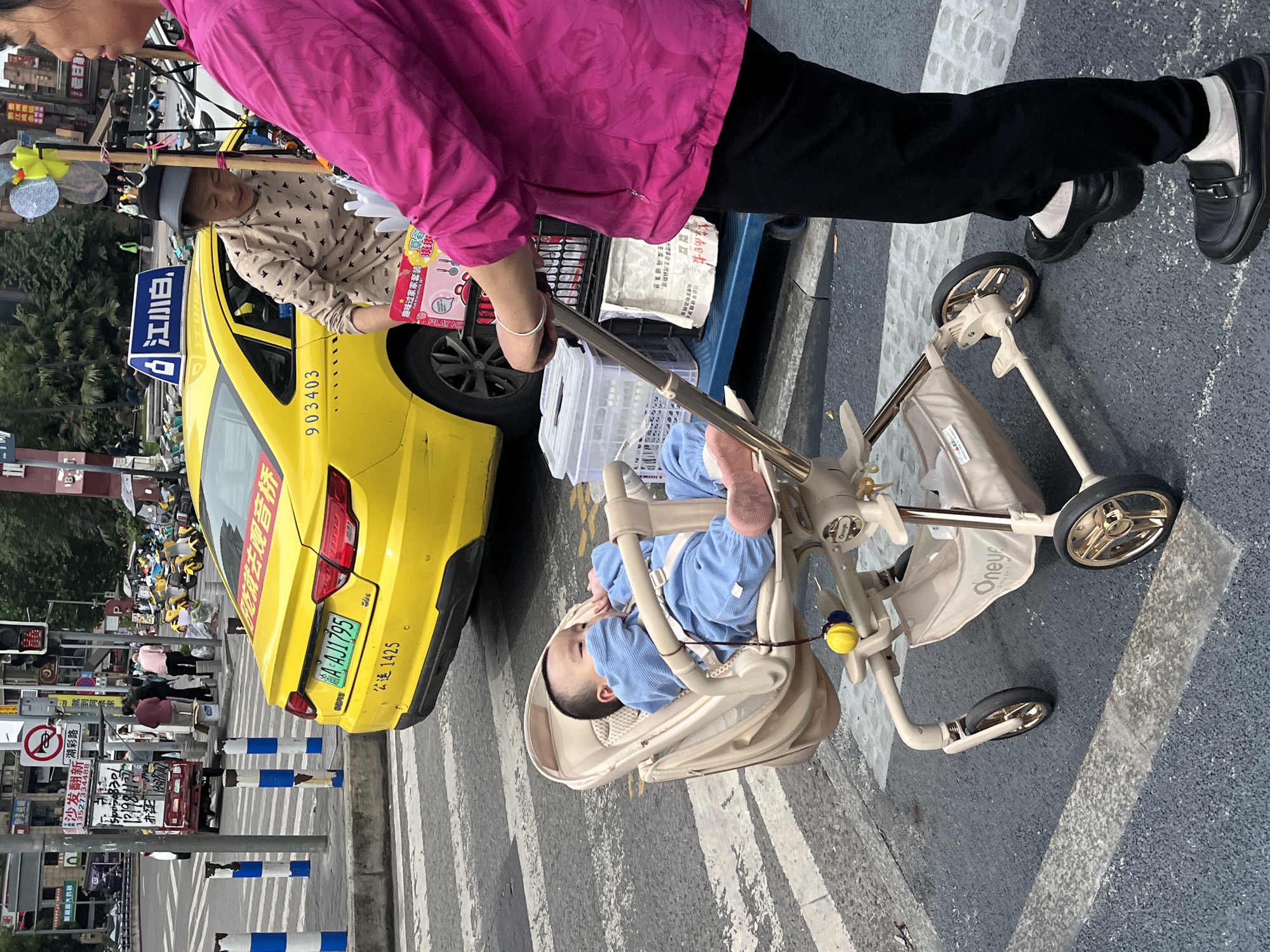 The image shows a street scene in Liangjiang, China. A baby in a tan stroller is being pushed across a pedestrian crosswalk. A person in a pink top and black pants pushes the stroller. A taxi is parked nearby. The license plate reads "A AJ1795." Signs in Chinese are visible on the taxi and surrounding area. The taxi number reads "903403." The name "江小白" is visible on a blue sign atop the taxi. A vendor stands near the taxi.