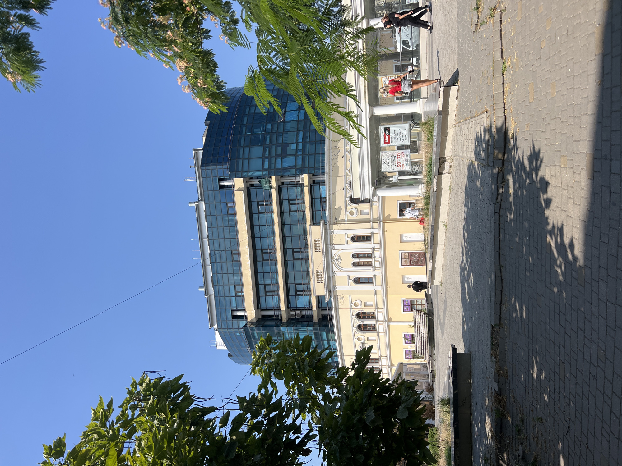 In Odesa, Ukraine, on a clear day with a bright blue sky, a modern glass-fronted building stands prominently behind a lower, pale yellow building with traditional architecture. The modern section features multiple floors of blue glass windows and balconies, contrasting with the arched windows and ornate detailing of the older structure. In the foreground, a paved walkway with visible shadows from overhead trees leads towards the buildings. Several people are present: a lone individual walks in the middle ground on the left side of the frame, while on the right, two women are walking away from the camera. One woman in a red top and denim shorts is in the process of sitting down on a low ledge. A white van is visible in the background to the right. Lush green trees with trailing seed pods frame the scene on the left, and blooming branches with pinkish flowers are seen on the right, suggesting a pleasant outdoor environment.