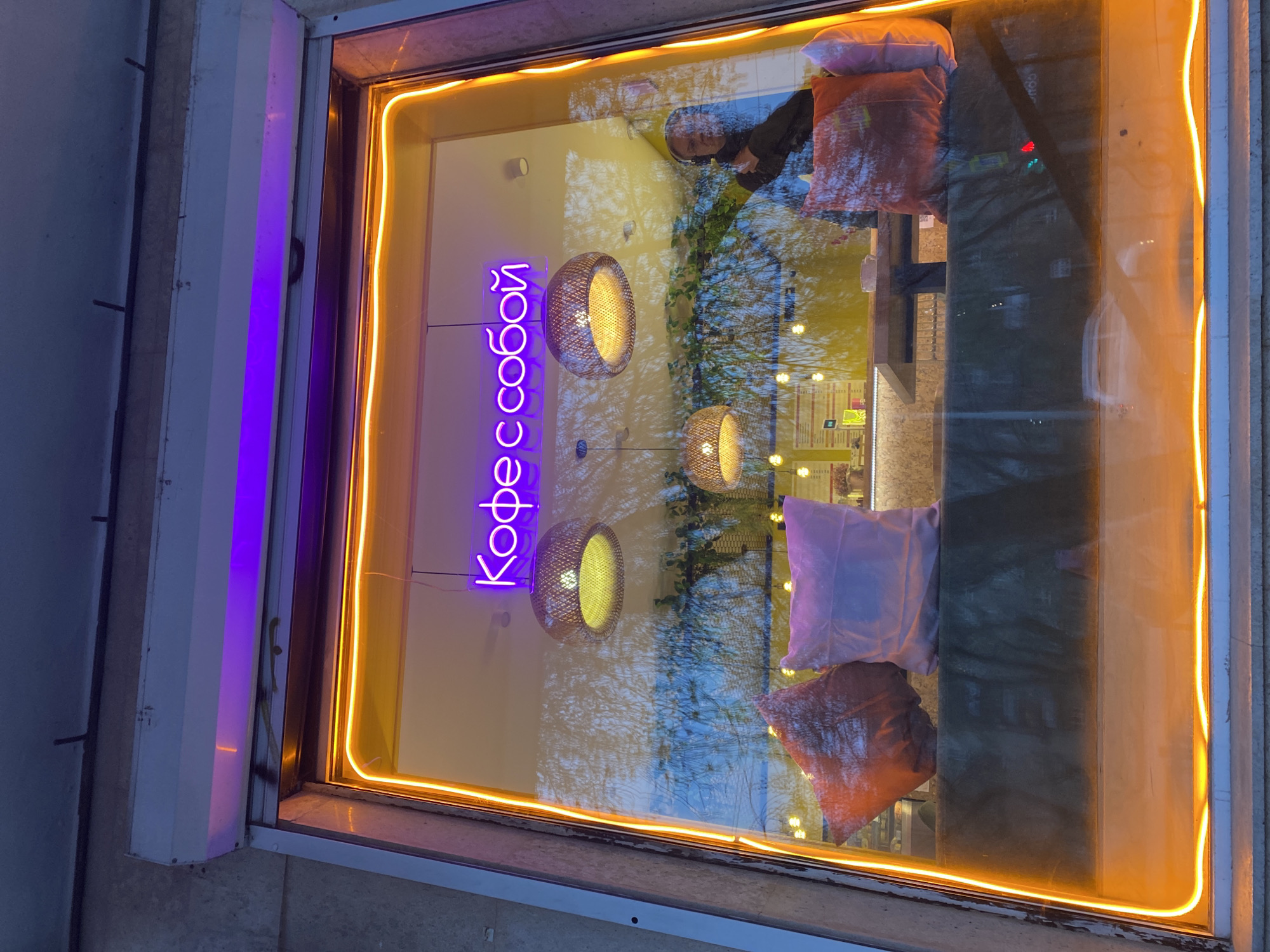 The image displays a storefront window, likely belonging to a café or coffee shop, viewed from the outside. The scene is illuminated by artificial lighting, suggesting dusk or nighttime.
Inside the window, a prominent purple neon sign spells out "Кофессобой" (Kofessoboy) in Cyrillic, which translates to "Coffee to go" or "Coffee with you." Below this sign, three circular pendant lights with a woven, possibly wicker or bamboo, texture hang, emitting a warm yellow glow. On what appears to be a window display or seating area, two decorative pillows are visible: one light purple and one reddish-brown. The entire window frame is outlined with a bright orange/yellow neon-like strip.
The glass of the window reflects the outside environment significantly. Clearly visible reflections include bare tree branches, some green foliage, and a pale blue sky, indicating an outdoor setting with natural elements. A reflection of a person with dark hair and clothing can be seen, possibly a passerby or someone inside looking out.
The combination of the vibrant neon signage, warm interior lights, and distinct reflections creates a dynamic visual. The Cyrillic text explicitly places the location in a Russian-speaking country, confirmed by the context to be Yekaterinburg, Russia.