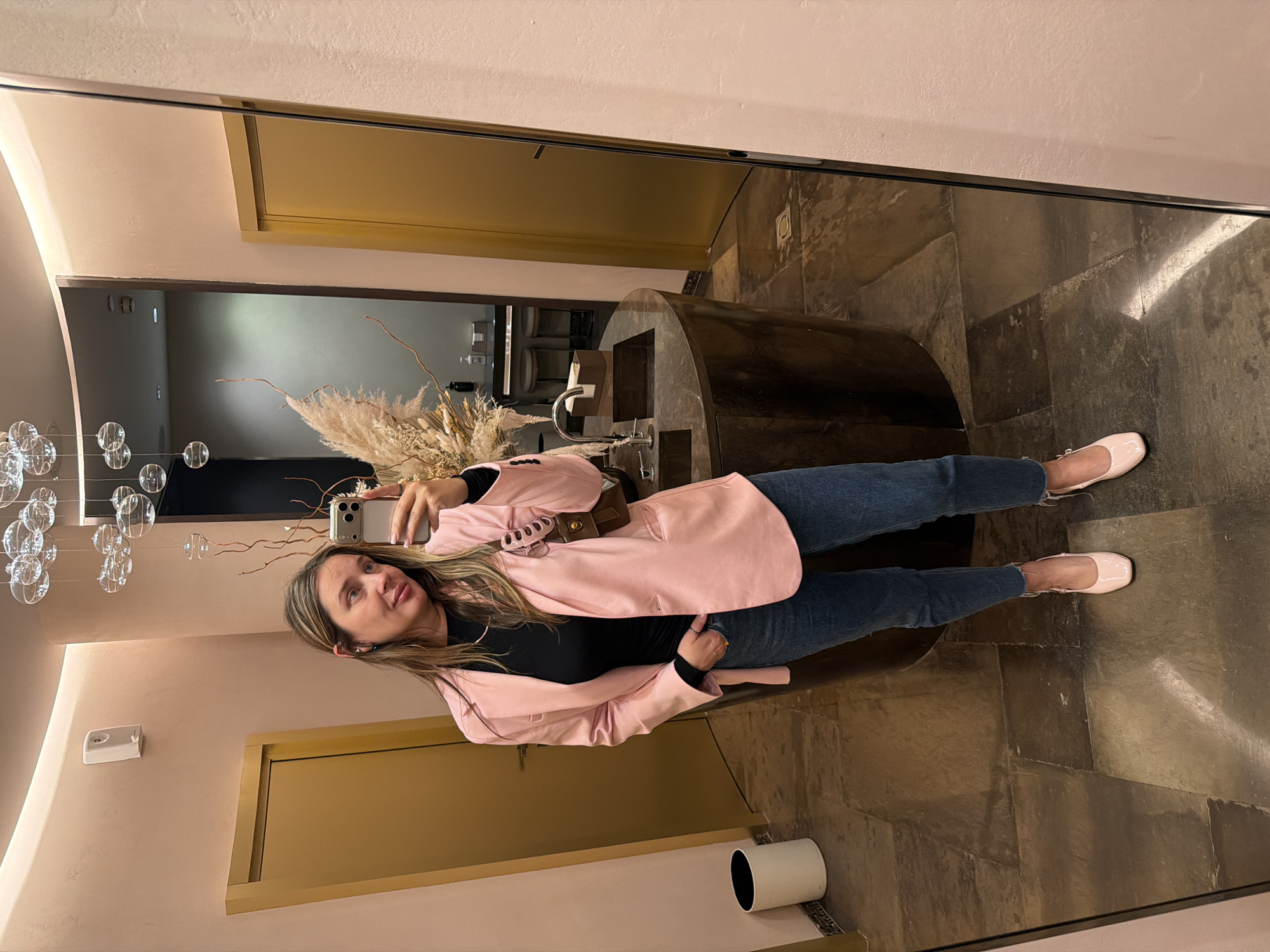 A woman stands in a room and takes a selfie in a mirror. She is wearing a pink blazer, a black top, blue jeans, and light pink ballet flats. Her hair is long and blonde, and she is wearing makeup. The room appears to be a hallway or an entrance area. To her left, there are decorative glass orbs hanging from wires, and to her right, there is a large, dark, curved object that might be a piece of furniture or a display. In the background, visible in the mirror, are doors and walls with a neutral color palette. The floor is tiled with large, dark stones. The overall impression is one of an indoor, possibly retail or public space, with a modern aesthetic. The lighting suggests it is daytime, but the interior setting makes it difficult to determine the exact time of day or weather conditions. There is no visible text in the image.