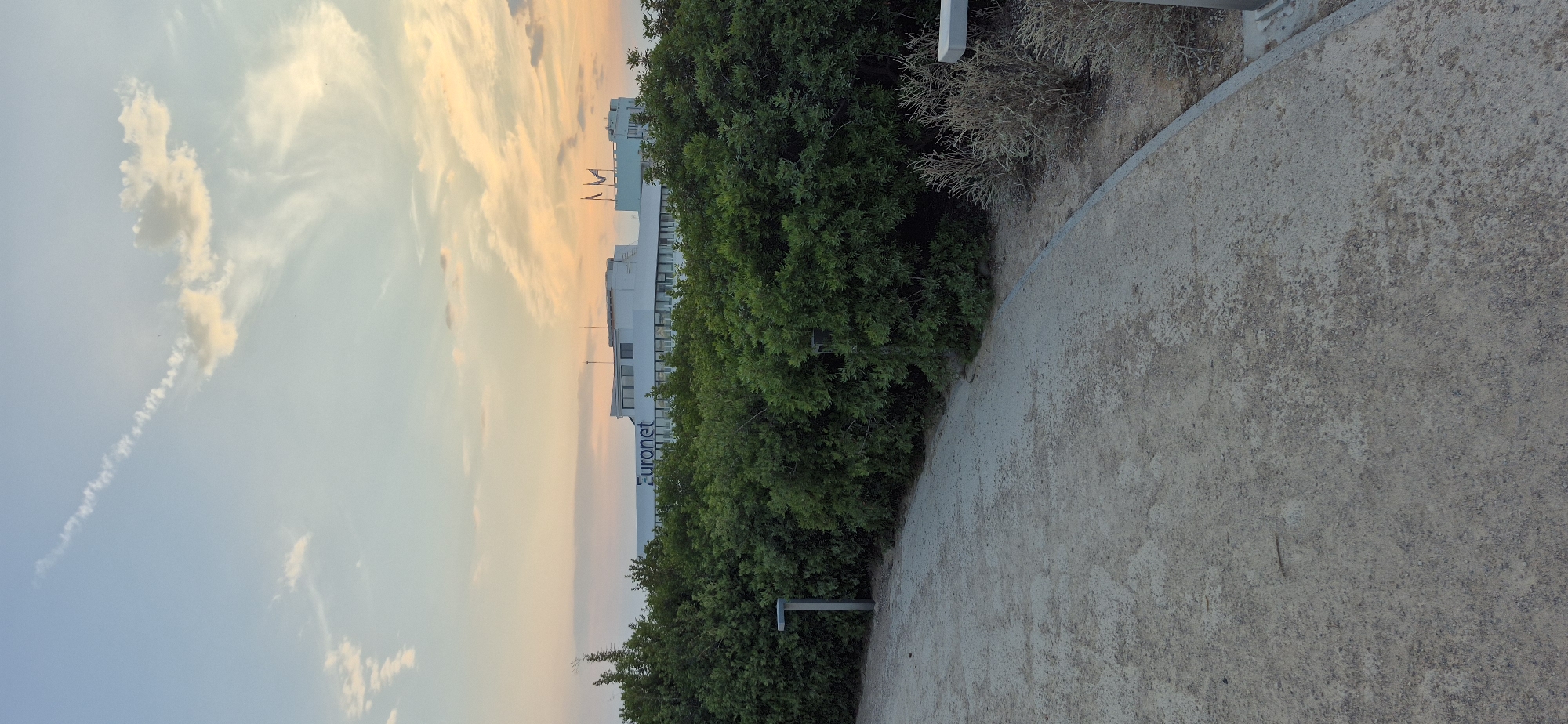 A slightly elevated view looks across a pathway and dense green bushes towards a white, modern building. The building features a horizontal sign that reads "Euronet". There are three flags mounted on poles on the roof of the building. The sky above is a soft blue with wispy clouds tinged with sunset colors of orange and pink. The pathway in the foreground appears to be made of light-colored gravel and is curving gently to the right. Two modern, grey outdoor lights are visible near the bushes. The overall scene suggests an outdoor area near an office building during a tranquil sunset.