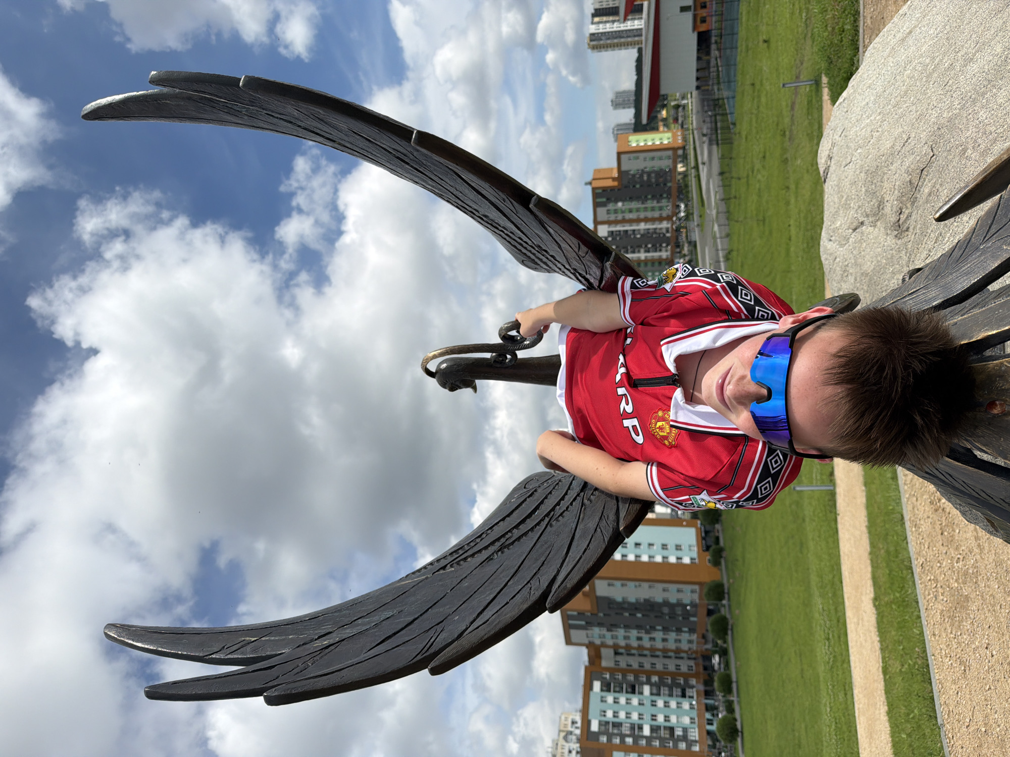 A young boy wearing a red Manchester United jersey and blue-tinted sunglasses is lying on a large bird statue with outstretched wings. The statue is dark and appears to be made of metal, with detailed feathering. The boy is resting his head on the statue's body, with his arm around its neck. He has short, brown hair and is looking towards the camera with a slight smile. The background shows a bright, cloudy sky and a city skyline with tall buildings. A grassy field and a paved pathway are also visible. The image is taken outdoors during the daytime, under a sky with scattered clouds, suggesting fair weather. The setting appears to be a public park or an urban area with a monument. The text visible on the jersey includes "SHARP" and "MANCHESTER UNITED", along with the club's crest. The location is Yekaterinburg, Russia.