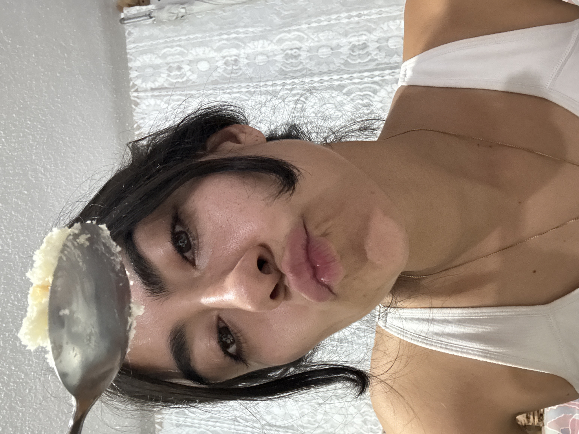 A young woman is holding a spoon of white dessert in front of her face. She is wearing a white top and a gold necklace. The background is a white lace curtain. There is no evidence in the photo to suggest a location in Puebla, Mexico.