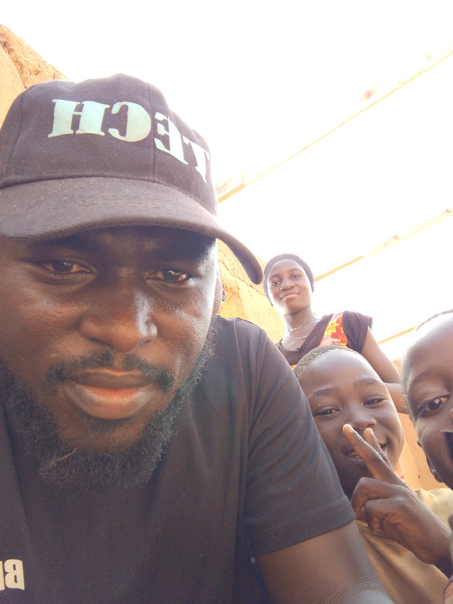 A selfie shows a dark-skinned man wearing a black "TECH" cap, black shirt, and a beard. Behind him are three children, two of whom are partially visible. One child is giving a peace sign. A young woman is standing further back. The location appears to be outside, in daylight, with a building or structure visible in the background. The city of Mulke Barikin Sayejo, Nigeria, is inferred from the prompt.