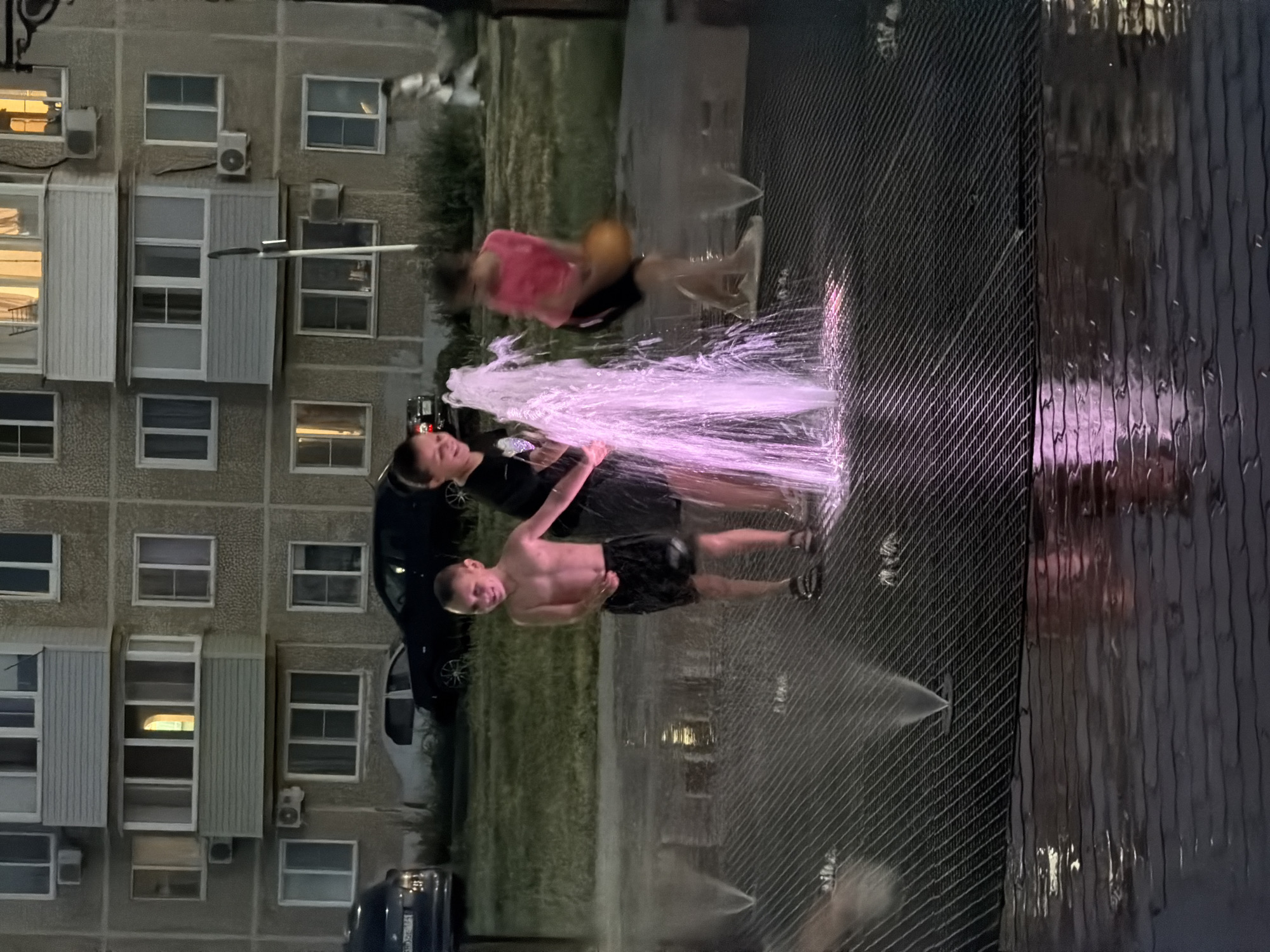 The image depicts an outdoor scene at dusk in Kamyshin, Russia, focusing on a group of children playing in a water fountain. In the foreground, a dark, textured surface, possibly a metal grate or paving, is wet and reflects the light. Water jets from the fountain are active, creating a pinkish glow. Three children are visible. One boy, bare-chested and wearing black shorts, stands near the center, smiling. To his left, another child, also seemingly a boy, in a black t-shirt and dark shorts, is positioned behind the fountain. To the right, a child in a pink t-shirt and black shorts is blurred, with an orange ball in their right hand. In the background, a multi-story residential building with numerous windows and balconies dominates the scene. Cars are parked in front of the building, and a patch of grass is visible. A person in white is walking in the far background on the right. The lighting suggests it is either early evening or late twilight.
