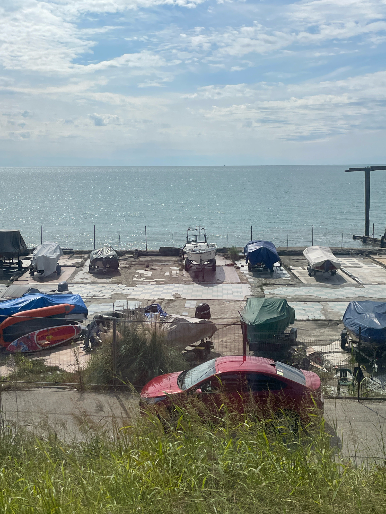 The image depicts a coastal scene with numerous boats covered with tarps, parked on a paved lot near the sea. The sea is calm and reflects the light, with a hazy sky above, suggesting a warm day. The boats are of various sizes and shapes, some on trailers. In the foreground, there's tall grass and a red car parked partially obscured by the vegetation. The scene appears to be an outdoor storage area for boats, possibly a marina or boatyard. There are no people visible in the image. The environment suggests a typical recreational area by the coast. This scene could be located in Sochi, Russia, known for its Black Sea coastline and numerous marinas and boat storage facilities.