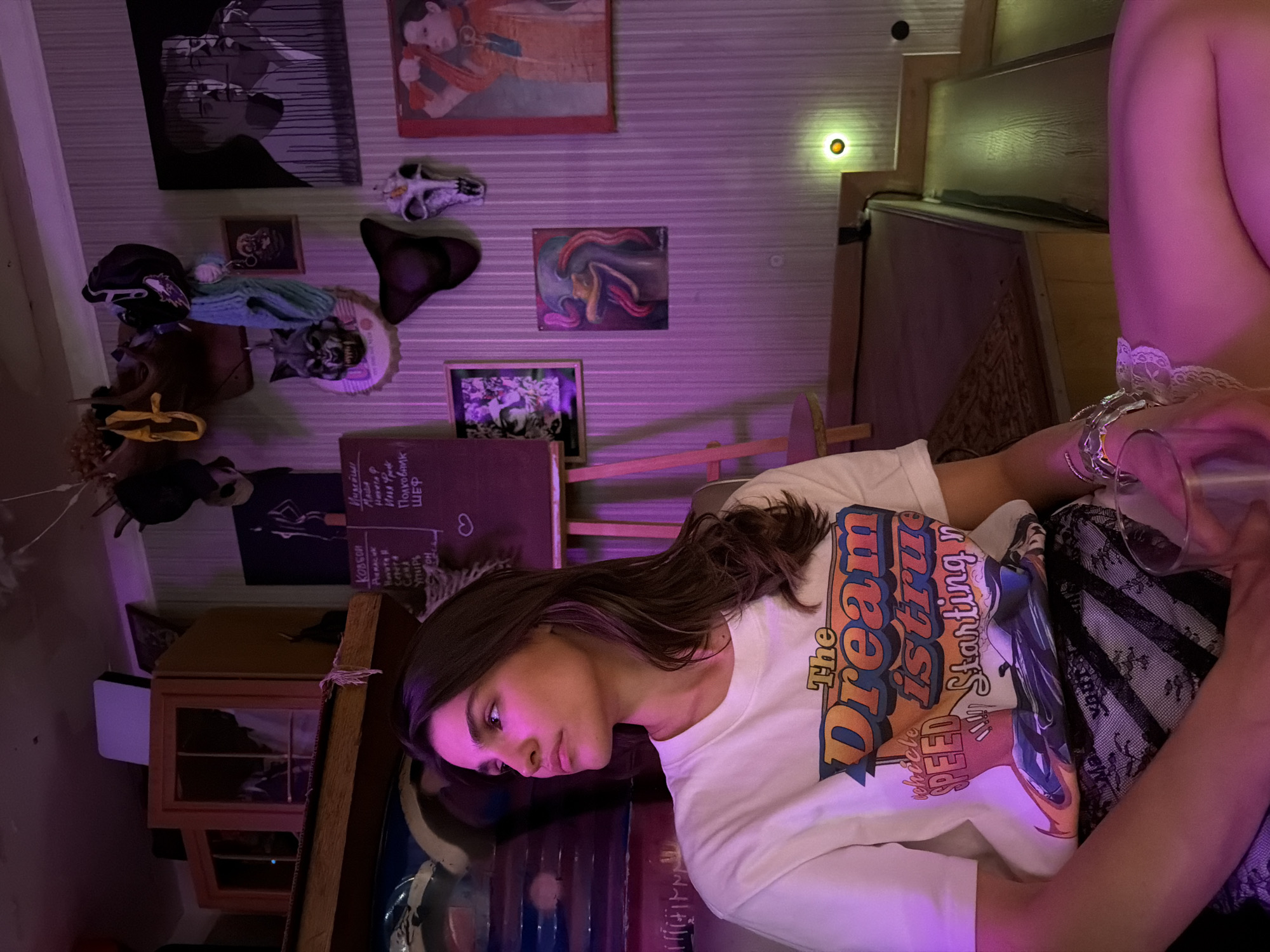 The image displays a young woman in an atmospheric, dimly lit indoor setting, possibly a bar or an eclectic room in Moscow, Russia. She is depicted from the chest up, looking downwards with a contemplative expression, holding a clear glass. She wears a white t-shirt adorned with a colorful graphic design that reads "The Dream Starting n" and "vehicle SPEED". A white lace garter is visible on her upper arm.
The room is illuminated by a predominant purple and pink ambient light, complemented by a warm yellow glow from a small wall fixture, suggesting an evening or nighttime setting. The walls are densely decorated with various artworks and objects, creating a bohemian aesthetic. Visible art includes a large black and white portrait, a painting of a figure in red and orange, and smaller framed pictures, one depicting a stylized horse-like creature and another with purple hues. Decorative items such as a skull, several hats (including a dark baseball cap and a fedora), and doll-like figures are also present. Wooden furniture, possibly part of a bar or shelving, frames the space.
A chalkboard or sign on the wall features several words, some in Cyrillic, providing location context. Text includes "AURACOK", "Camera", "Cent", "Упырь" (Vampire), "OH", "Индейци" (Indians), "MALA", "Havera P", "Илья Фреск" (Ilya Fresco), "Полковник" (Colonel), and "ШЕФ" (Chief). The scene evokes a relaxed, artistic, and unique ambiance.