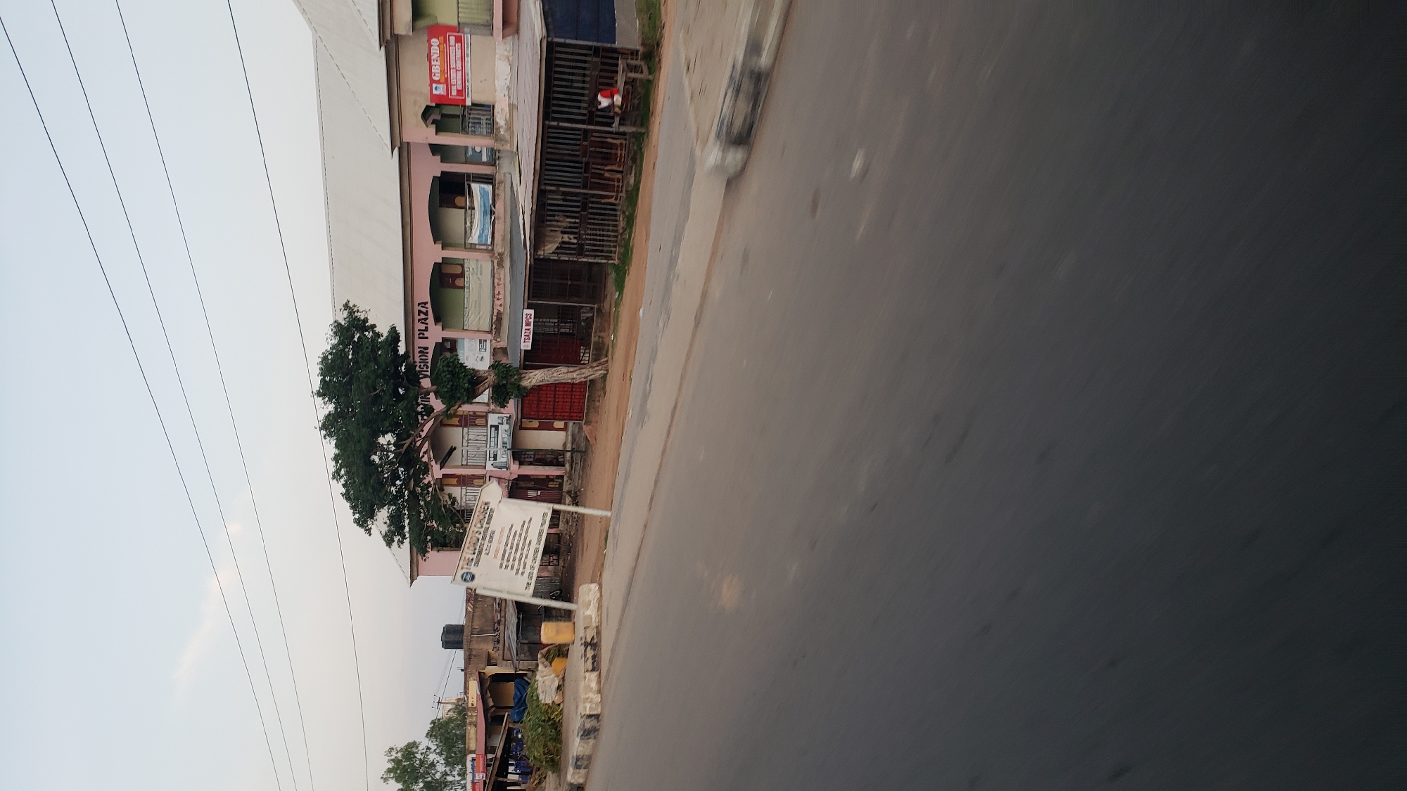 The image captures a street scene in Jalingo, Nigeria, with a prominent pink building labeled "VISION PLAZA". A large tree stands in front of the building, obscuring parts of its facade. To the left of the plaza, a sign for "THE LORD'S CHOSEN CHARISMATIC REVIVAL MINISTRIES" is visible. Adjacent to the plaza are several stalls with metal bars, suggesting commercial activity. In the foreground, a road with visible lane markings indicates it is a public thoroughfare. A person in a red shirt is seated at one of the stalls on the right side of the image. The sky above is clear, and power lines are strung across the scene.