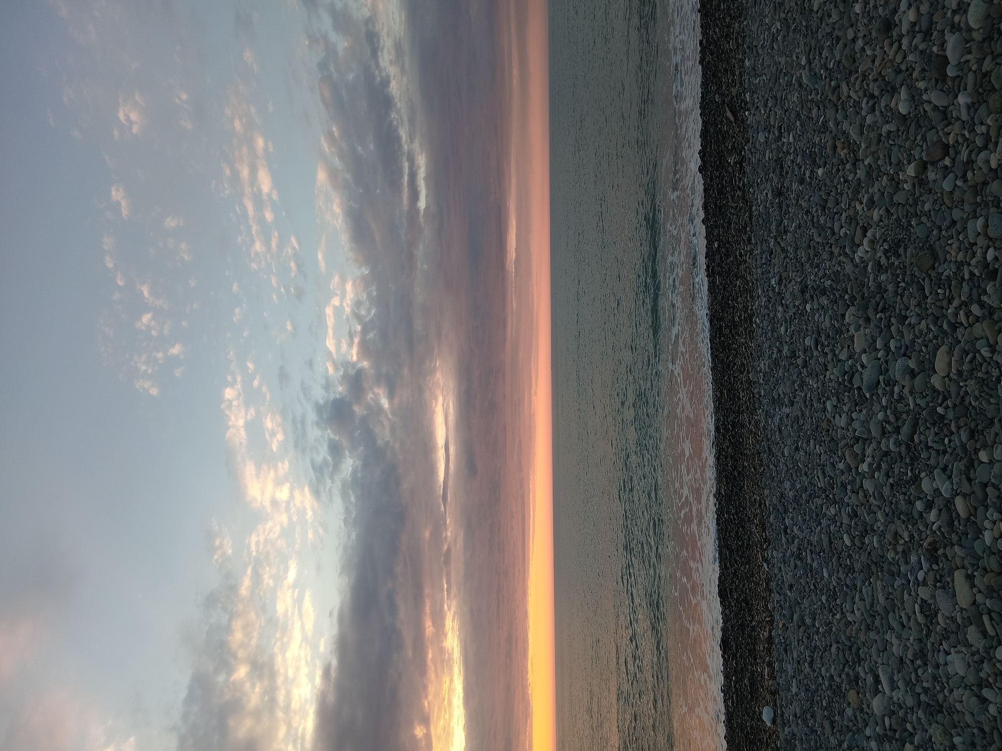 The image depicts a view of the Black Sea coast at sunset in Batumi, Georgia. The foreground shows a pebble beach with small, dark grey stones. The sea is calm with small waves washing onto the shore. The sky is filled with clouds that are illuminated by the setting sun, with colors ranging from pale blue to orange and pink. There are no people visible in the scene.