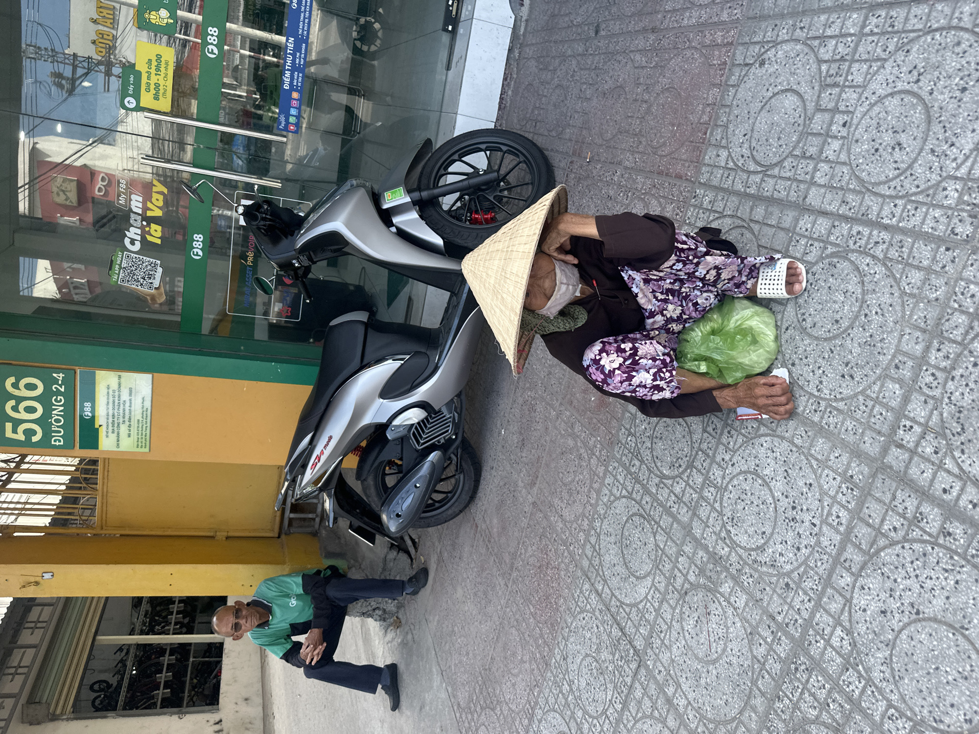 In Bac Nha Trang Ward, Vietnam, a street scene unfolds with two individuals in the foreground. To the left, a seated man, appearing elderly with a bald head and glasses, is dressed in a green and black polo shirt and dark pants. He sits on a low concrete ledge, hands clasped. In front of him rests a silver scooter. To the right and closer to the camera, an elderly woman, wearing a traditional conical hat, a face mask, and a purple floral patterned garment, is seated on the tiled sidewalk. She holds a green plastic bag and rests her chin on her hand, looking down. The background features a yellow building with signage, a glass-fronted storefront displaying merchandise and advertisements, and more scooters. The ground is paved with patterned tiles.