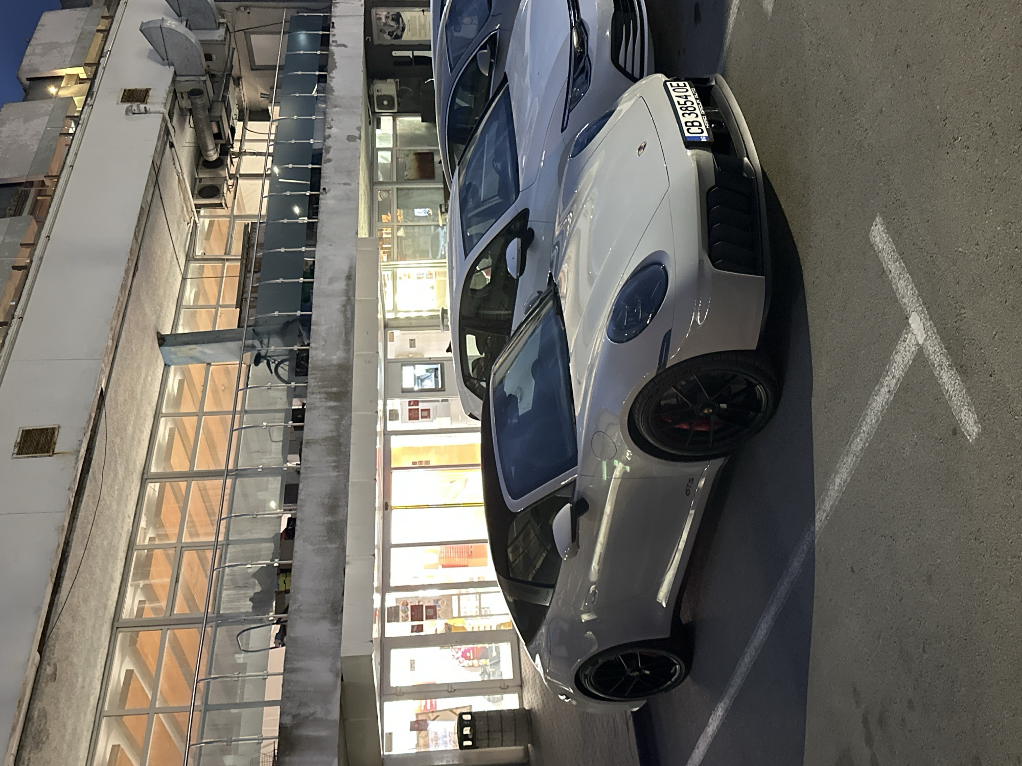 The image shows a white Porsche GTS parked in front of a building in Varna, Bulgaria. The car's license plate is "CB 3854 OE." The building appears to be a commercial structure with multiple floors and large windows. The lighting suggests it might be evening or early morning. The parking space is marked with white lines on the asphalt surface. Another vehicle is partially visible next to the Porsche.