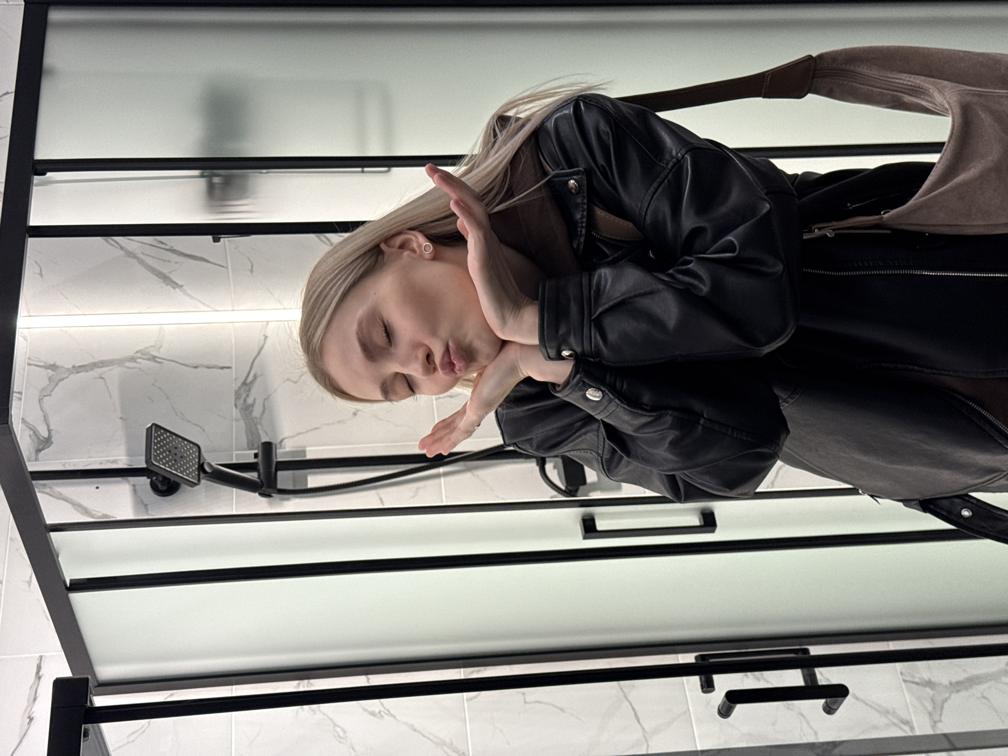 A blond woman in a black leather jacket stands in a bathroom. The bathroom features white marble-patterned tiles, a black-framed shower, and a shower head. A beige bag is slung over her shoulder. The woman is posed with her eyes closed and her hands near her face, in what appears to be a kissing gesture. The location is unknown and not inferable from the image.