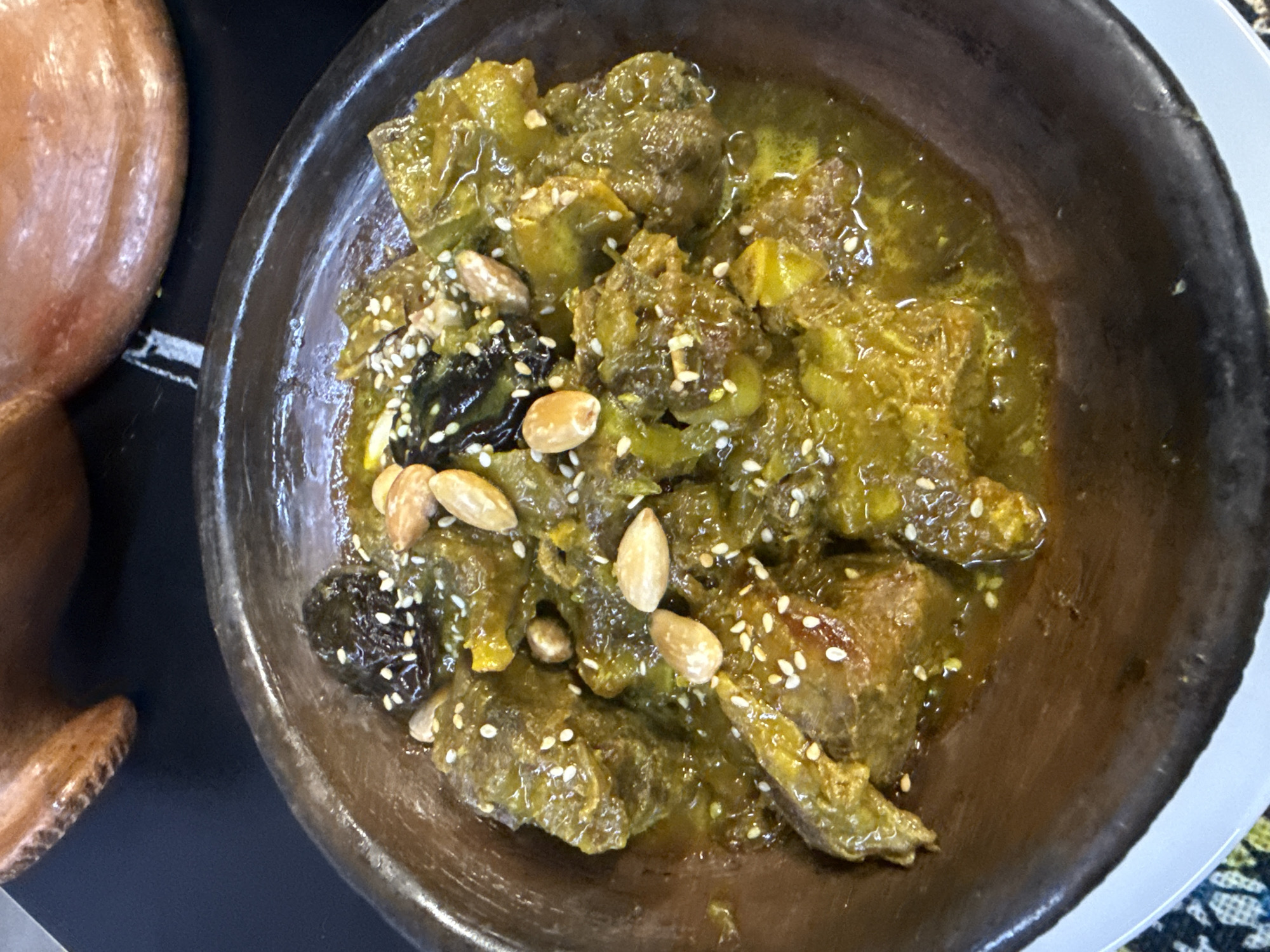 A close-up, high-angle shot captures a rustic bowl filled with a rich, savory stew, presented in what is contextually identified as Barcelona, Spain. The dish, possibly a tagine, features tender-looking chunks of meat and green vegetables, all bathed in a thick, yellowish-green sauce that suggests a blend of spices and possibly preserved lemons or artichokes. The surface of the stew is generously garnished with whole blanched almonds, scattered white sesame seeds, and dark, wrinkled pieces of dried fruit, resembling prunes or dates, adding a mix of textures and flavors.
The food is served in a substantial, dark brown, and seemingly handcrafted ceramic or clay bowl with a thick, uneven rim, enhancing its traditional appearance. Part of another light brown, rounded pottery item is visible on the far left, suggesting a complementary piece of dinnerware. The bowl rests on a dark surface, possibly a table, with a glimpse of a lighter-colored plate or cloth and a subtly patterned fabric peeking from the bottom and right edges of the frame. There are no people visible in the image, and no discernible text is present.