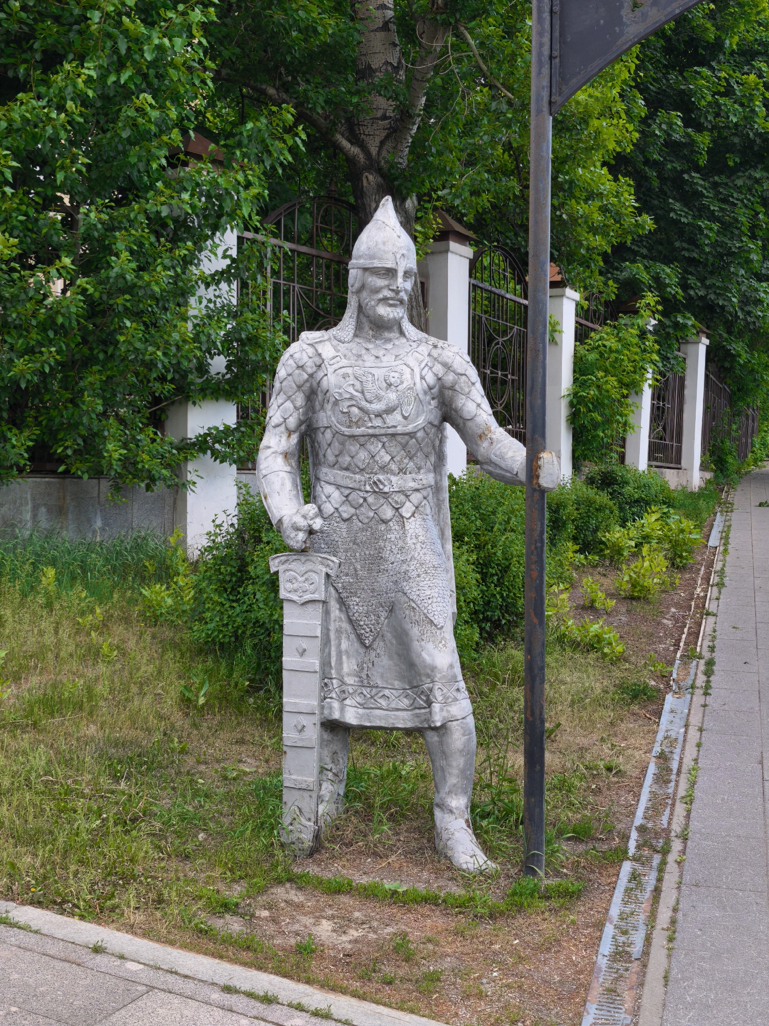 A full shot captures a stone statue of a medieval knight in front of a green background. The knight stands on a patch of grass beside a paved sidewalk. The statue is gray, weathered, and detailed with chainmail and armor. He wears a pointed helmet and holds a sword. Behind the statue is a metal fence, white concrete posts, and dense green foliage. To the right of the statue is a rusty metal pole. The sidewalk runs along the edge of the frame. Based on the statue and general appearance, the location is likely in Moscow, Russia.