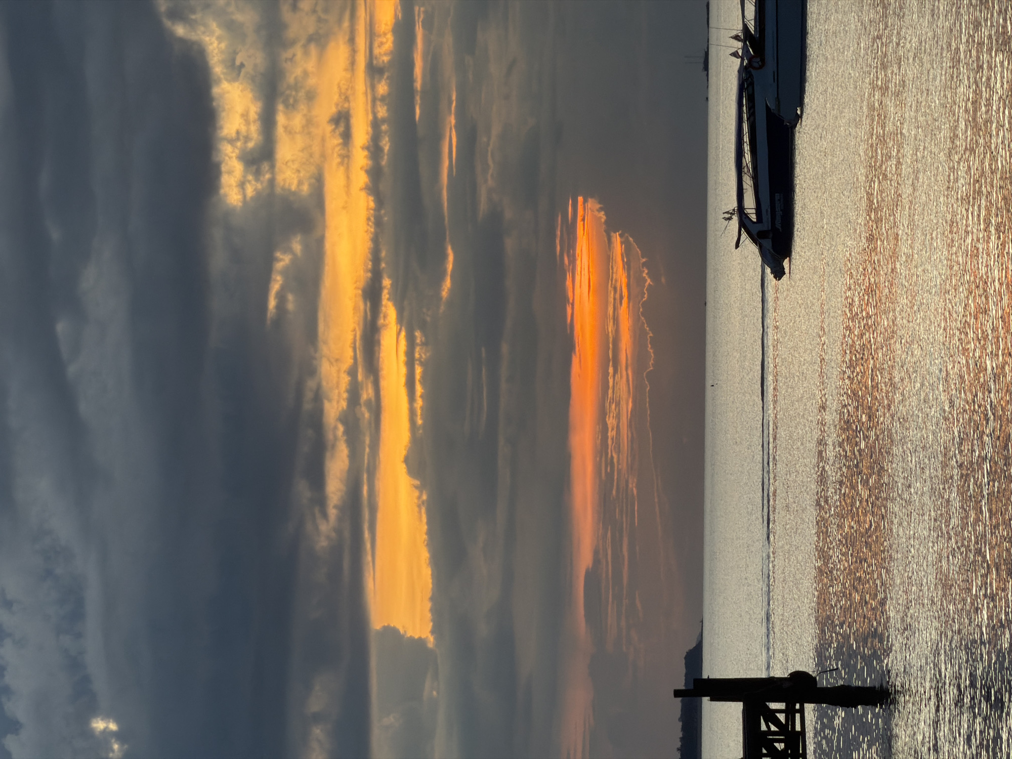 A sunset over the ocean in Baan Bang Rak, Thailand, features dramatic clouds in shades of gray and orange. The water reflects the sky's colors, creating a shimmering surface. In the foreground, a wooden pier juts out into the water, with its pilings silhouetted against the sea. Two speedboats are moored in the middle ground, their sleek designs contrasting with the natural elements. In the background, a faint landmass is visible on the horizon, suggesting the proximity of the coastline. The overall scene evokes a sense of tranquility and natural beauty, with the vibrant sunset painting the sky and sea in warm hues.