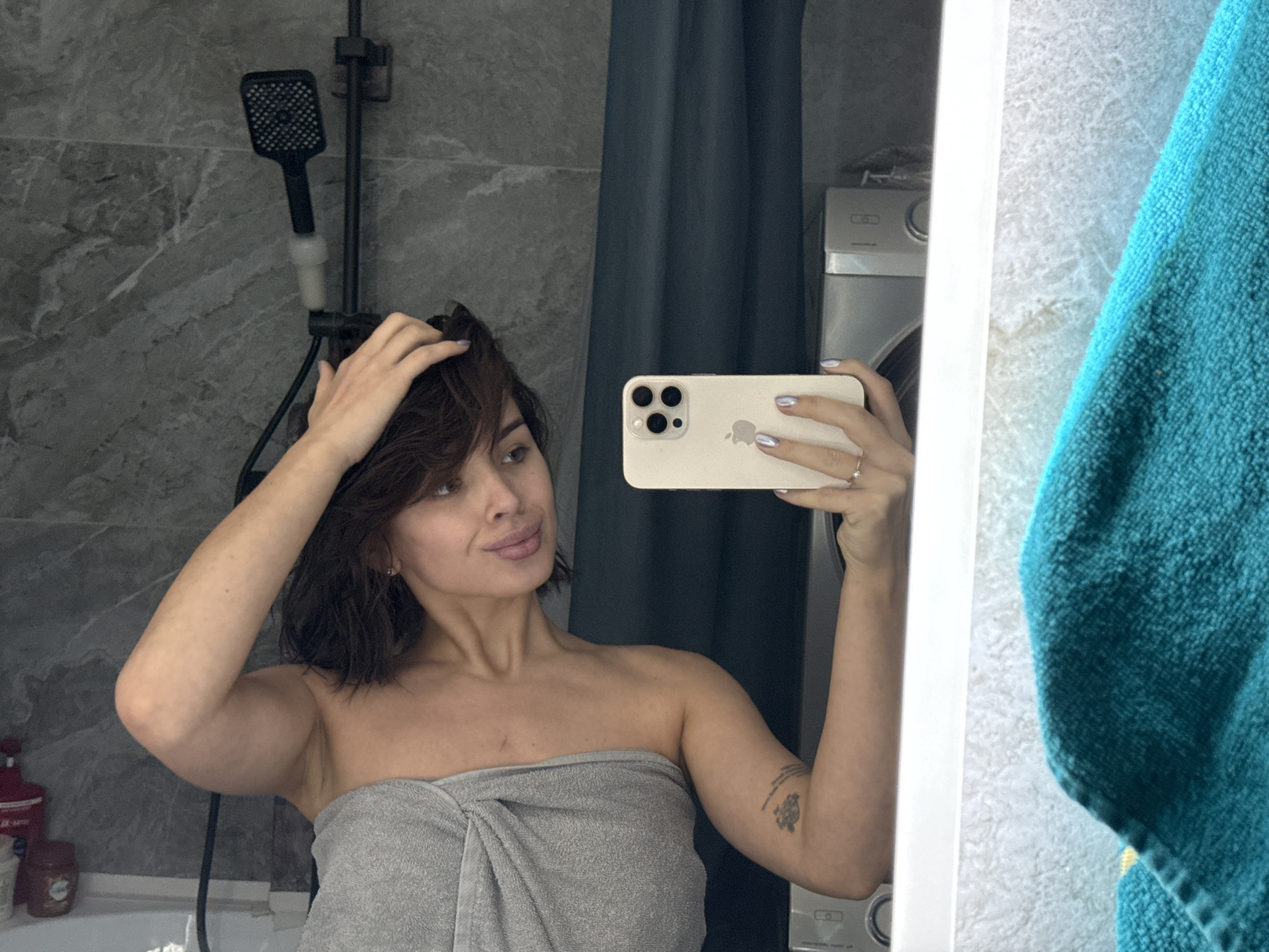A woman stands in a bathroom, holding a smartphone in her right hand, taking a selfie in the mirror. She is wrapped in a gray towel. Her dark hair is slightly wet, and she is touching it with her left hand. A showerhead is visible on the marble-patterned wall behind her. A washing machine is visible in the background to the right. A blue-green towel hangs partially in front of the mirror on the right side. Based on the bathroom interior and the washing machine, the location is likely a private residence in the city of Лобаново, Russia.