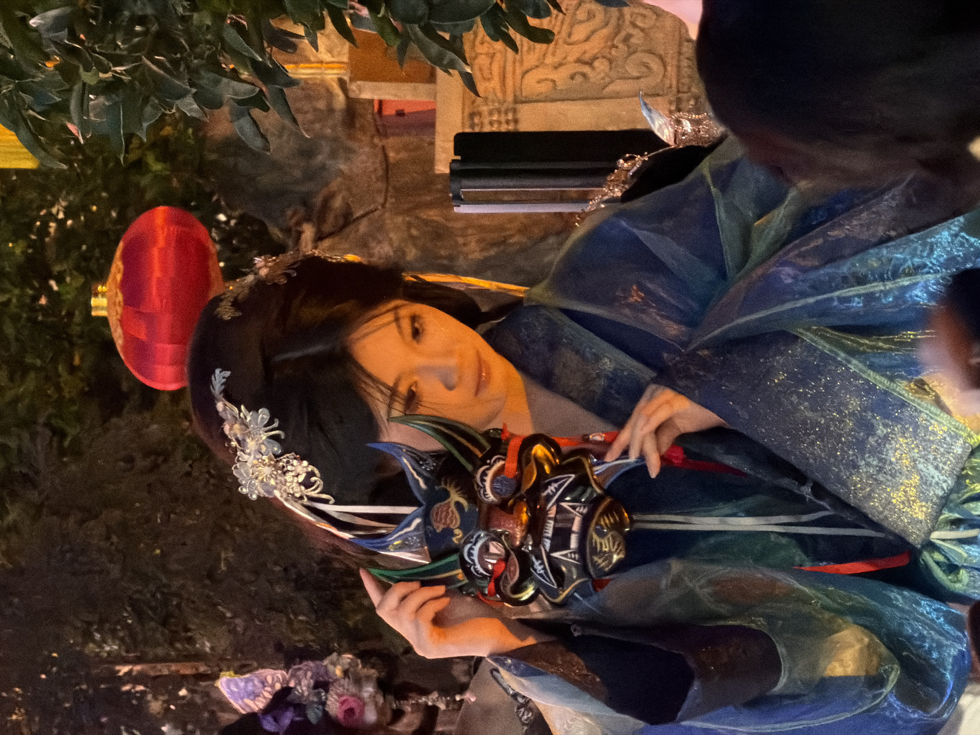 The image shows a person dressed in traditional Chinese attire. The person has dark hair styled with an ornate silver hairpiece. They are wearing a layered blue and green robe with gold detailing, and a decorative mask with intricate designs and red ribbons. The person is holding the mask with their hands. The background is dimly lit, suggesting it is nighttime or indoors with artificial lighting. There are some green leaves and a red lantern visible on the left side of the image. The setting appears to be an outdoor or semi-outdoor event, possibly a festival or performance, given the attire and the presence of decorative elements. The person's gaze is directed downwards towards the mask, and their expression is somewhat contemplative. The overall mood is one of traditional beauty and cultural display. There is no visible text in the image.