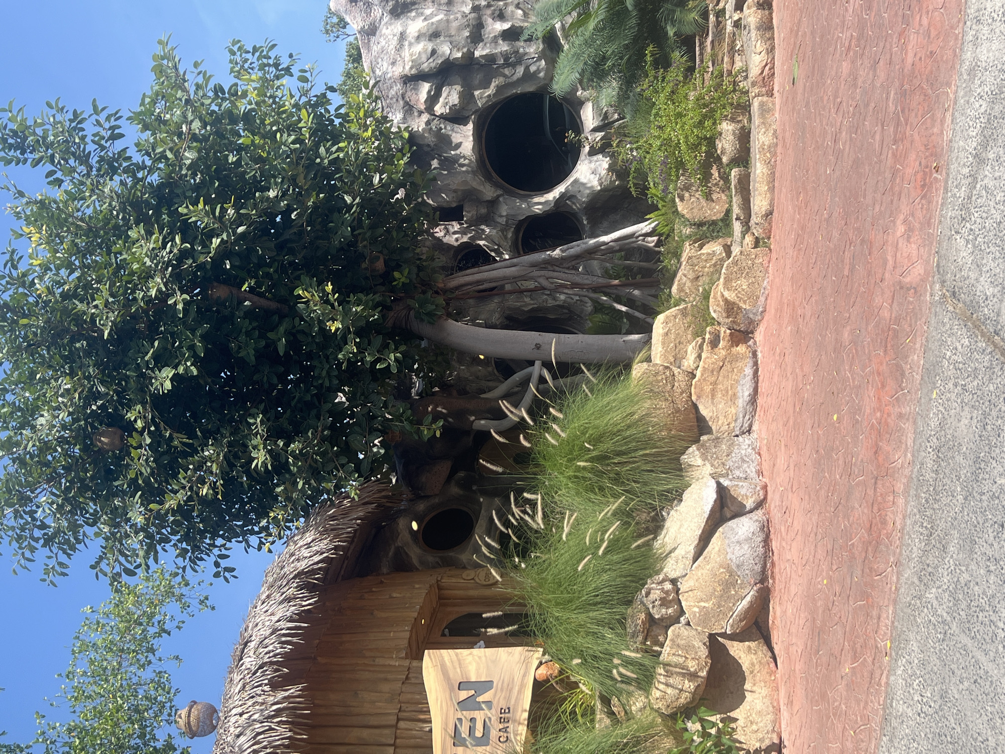 The image depicts a whimsical and naturalistic outdoor setting, likely a cafe, on the island of Ko Samui, Thailand. The architecture features a thatched-roof structure made of wood, with a prominent sign that reads "TEN CAFE." Adjacent to this is a rock formation designed to resemble a cave or a natural cliff face, with several circular openings that serve as windows. A large tree with exposed roots grows in front of the rock formation, and its branches extend over the thatched roof. The ground is paved with reddish-orange interlocking bricks, and large rocks and lush green vegetation, including tall grasses, are arranged around the entrance. The sky above is clear and blue, indicating a sunny day. There are no people visible in the frame. The overall impression is one of a creative and nature-integrated establishment, possibly designed to mimic a hobbit hole or a natural dwelling.