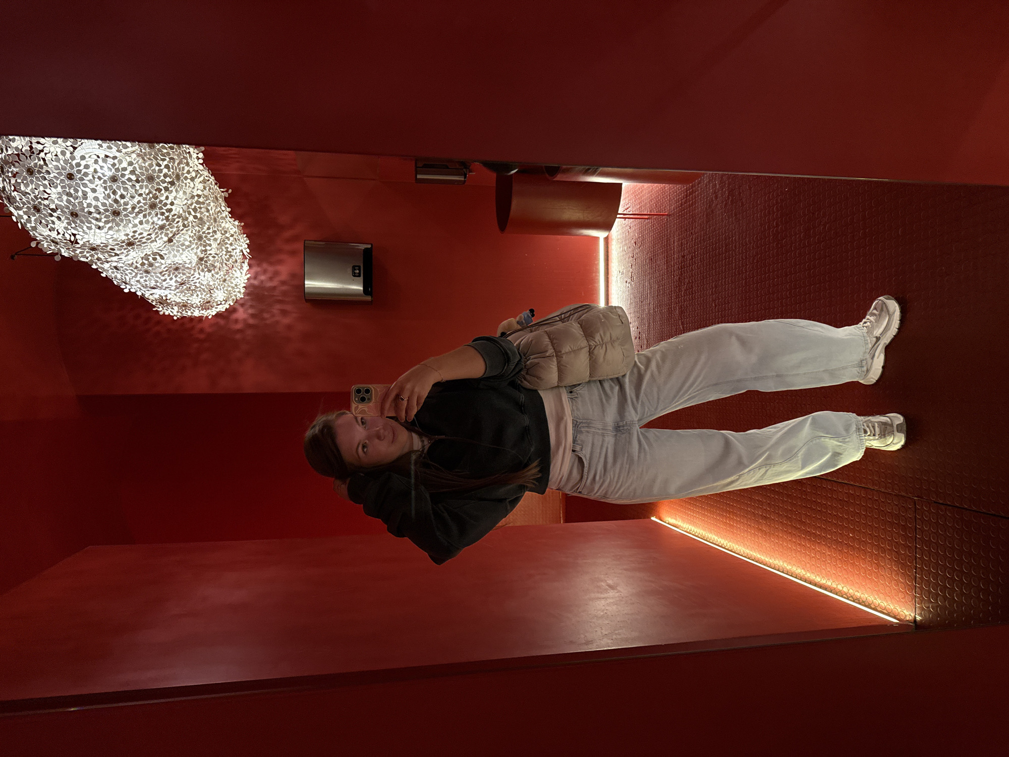 A young woman captures a mirror selfie in a strikingly red-themed indoor space. She stands upright, holding a smartphone with a light-colored case up to her face, looking directly at the camera with a slight smile. She has long brown hair and is dressed casually in a dark long-sleeved top, light-wash jeans, and a beige puffer vest. A beige quilted shoulder bag is slung over her shoulder, and she wears white athletic shoes, one of which prominently displays the Nike swoosh logo.
The room is characterized by its intense red color scheme. The floor has a textured pattern of small, overlapping circles. Along the base of one wall, a linear light strip emits an orange-red glow, highlighting the floor's texture. On an adjacent red wall, a large, intricate light fixture resembling a cloud or cluster of white, petal-like elements hangs, casting complex shadows. Below this decorative light, a silver rectangular object, possibly a hand dryer or dispenser, is mounted on the wall. Another curved, red architectural detail is visible further back in the reflection.
The setting appears to be a modern and distinctive interior, possibly a public space like a restroom or a gallery within a building in Belgrade, Serbia, given its artistic lighting and bold color choice. The time of day is not discernible due to the artificial lighting. No other text is visible besides the Nike logo.