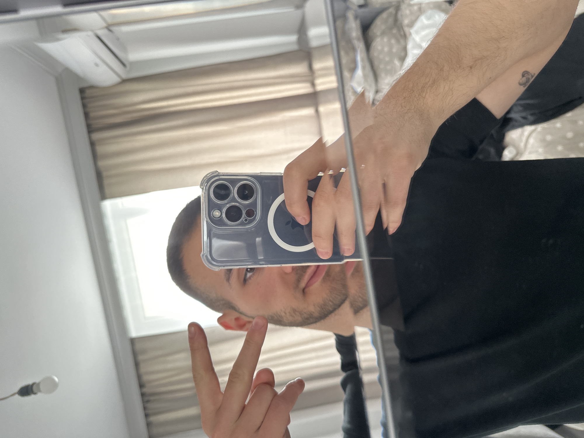 A man is taking a mirror selfie in what appears to be a bedroom. He is wearing a black t-shirt and making a peace sign with his left hand. In his right hand, he is holding a smartphone, which is being used to take the photo. The background shows curtains, an air conditioning unit, and a bed with patterned bedding. A bare lightbulb hangs from the ceiling. There is a small tattoo of what appears to be a crescent moon with a star on the man's left forearm. This scene does not provide any specific visual cues to determine the location within the city of Varna, Bulgaria.