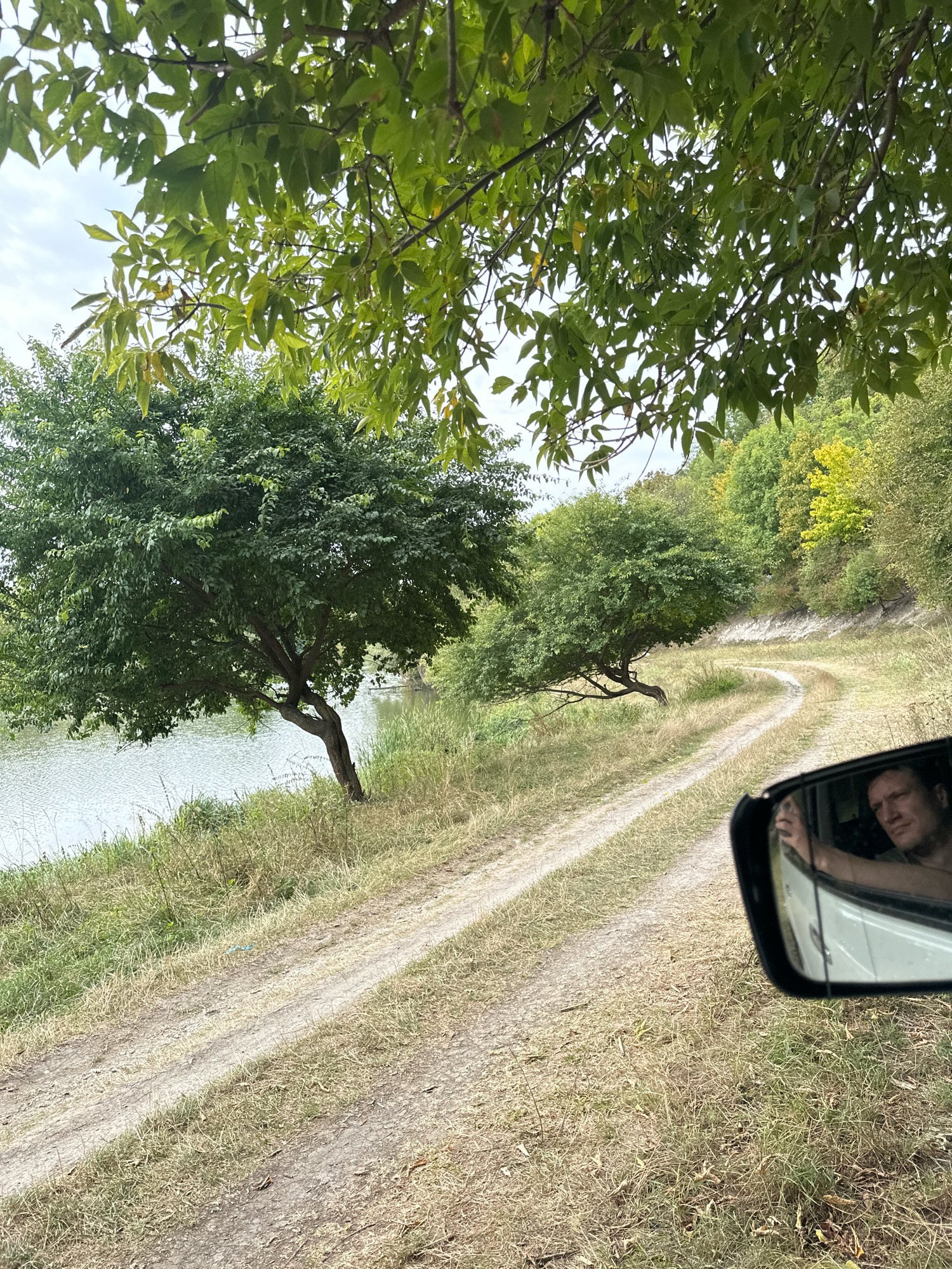 The image depicts a dirt road winding through a natural landscape, likely in or near Makiv, Ukraine. On the left, a body of water, possibly a river or lake, is visible, bordered by dry grass and a large, leafy tree. Beyond the water, a lush green hillside is dotted with trees, some exhibiting yellowing leaves, suggesting an autumn season. The road itself is rutted, indicating it is frequently used by vehicles. In the foreground, a side-view mirror of a white vehicle reflects the face of a man with fair skin and short, dark hair. He is looking forward, his left arm resting on the open window frame. The sky is overcast.