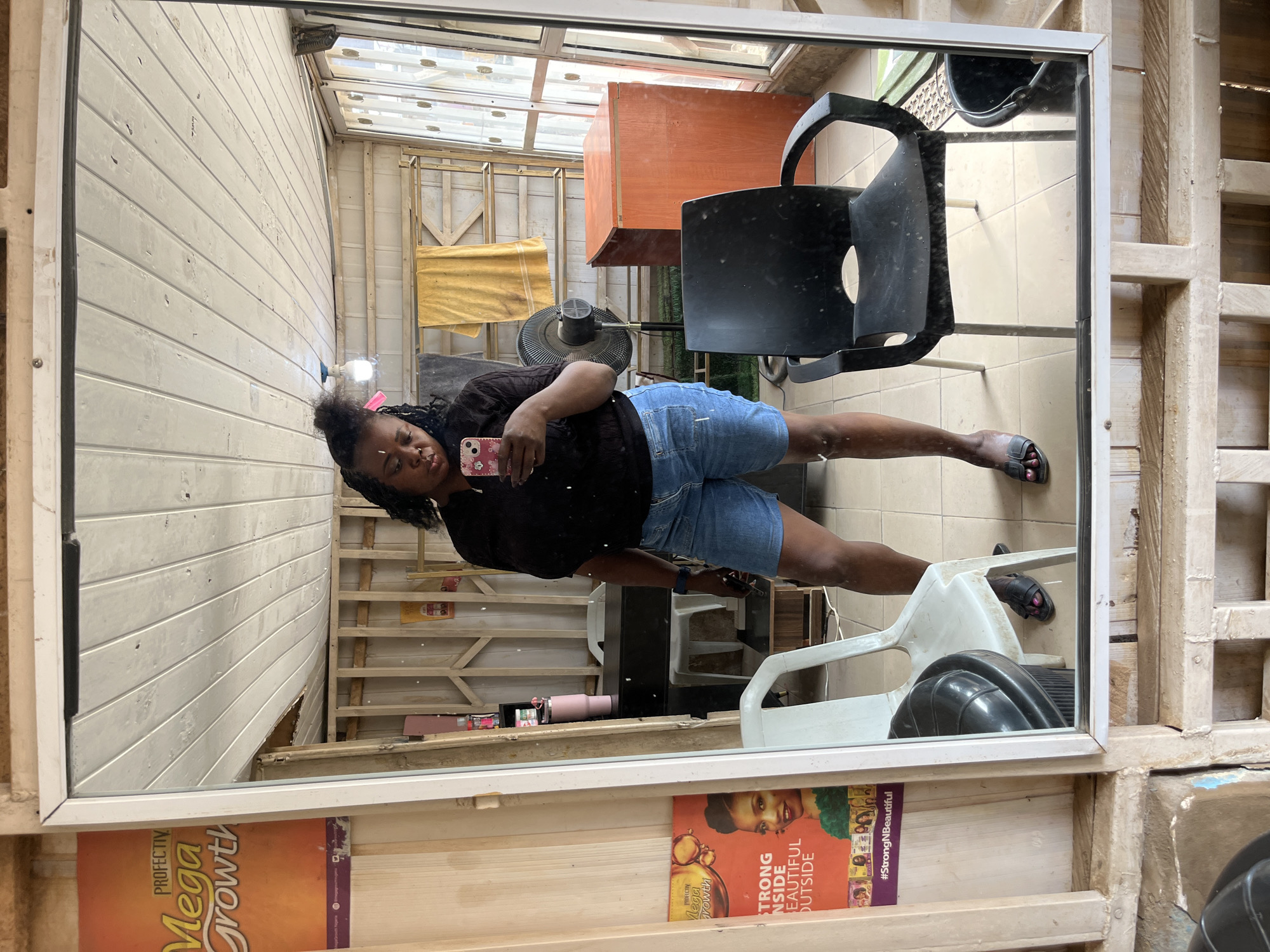 This media file is a mirror selfie taken by a Black woman in what appears to be a salon or workshop in Abuja, Nigeria. The woman is wearing a black t-shirt and denim shorts, and she is holding a pink-cased smartphone. She has braided hair and is wearing black sandals.
The setting is rough and unfinished, with exposed wooden beams and corrugated metal walls visible in the reflection. A black salon chair is in the foreground, and a white plastic chair is behind it. In the background, there is a small orange cabinet and a fan. There is also a poster on the wall behind the woman with text that is partially visible.
The lighting suggests it could be daytime, possibly with natural light coming from overhead. The overall mood seems casual and candid, with the woman seemingly relaxed as she takes the photo.
Visible text on the poster in the background includes "Mega Growth" and "#StrongNBeautiful". The poster also features images of women with diverse hairstyles.