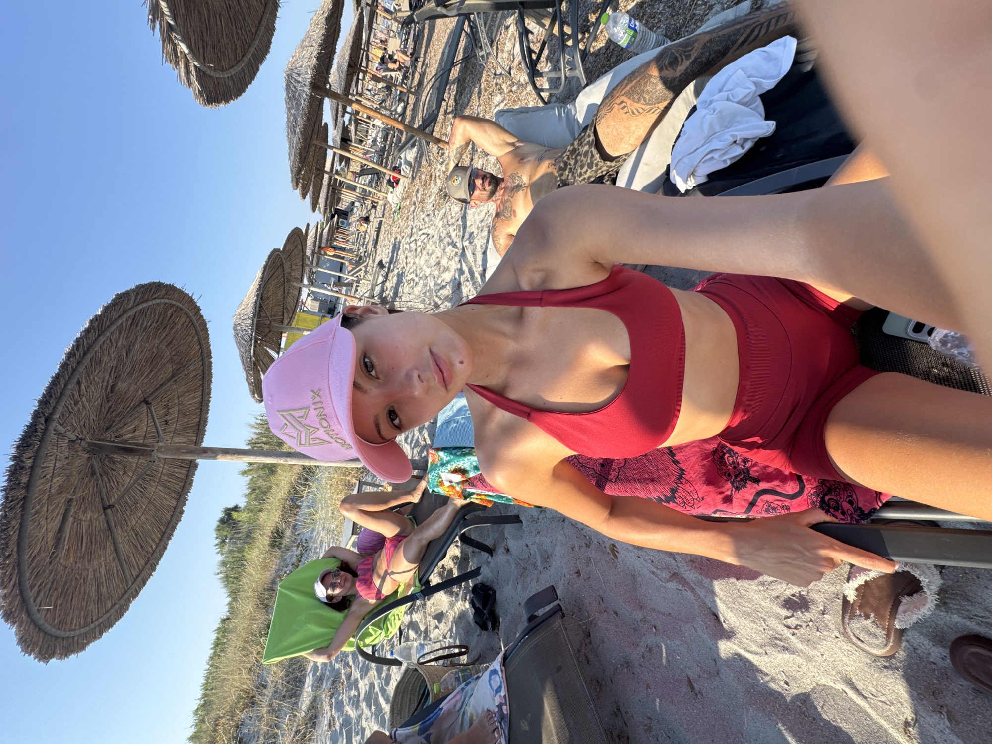 A group of people are relaxing on a sandy beach in Moudros Municipal Unit, Greece, under thatched umbrellas. In the foreground, a woman wearing a red bikini and a pink cap takes a selfie. Behind her, another woman in a pink bikini reclines on a lounge chair. To her right, a tattooed man in patterned shorts is also lounging. Several beach chairs and umbrellas are visible in the background, along with distant figures and sparse vegetation. The weather appears sunny and clear.