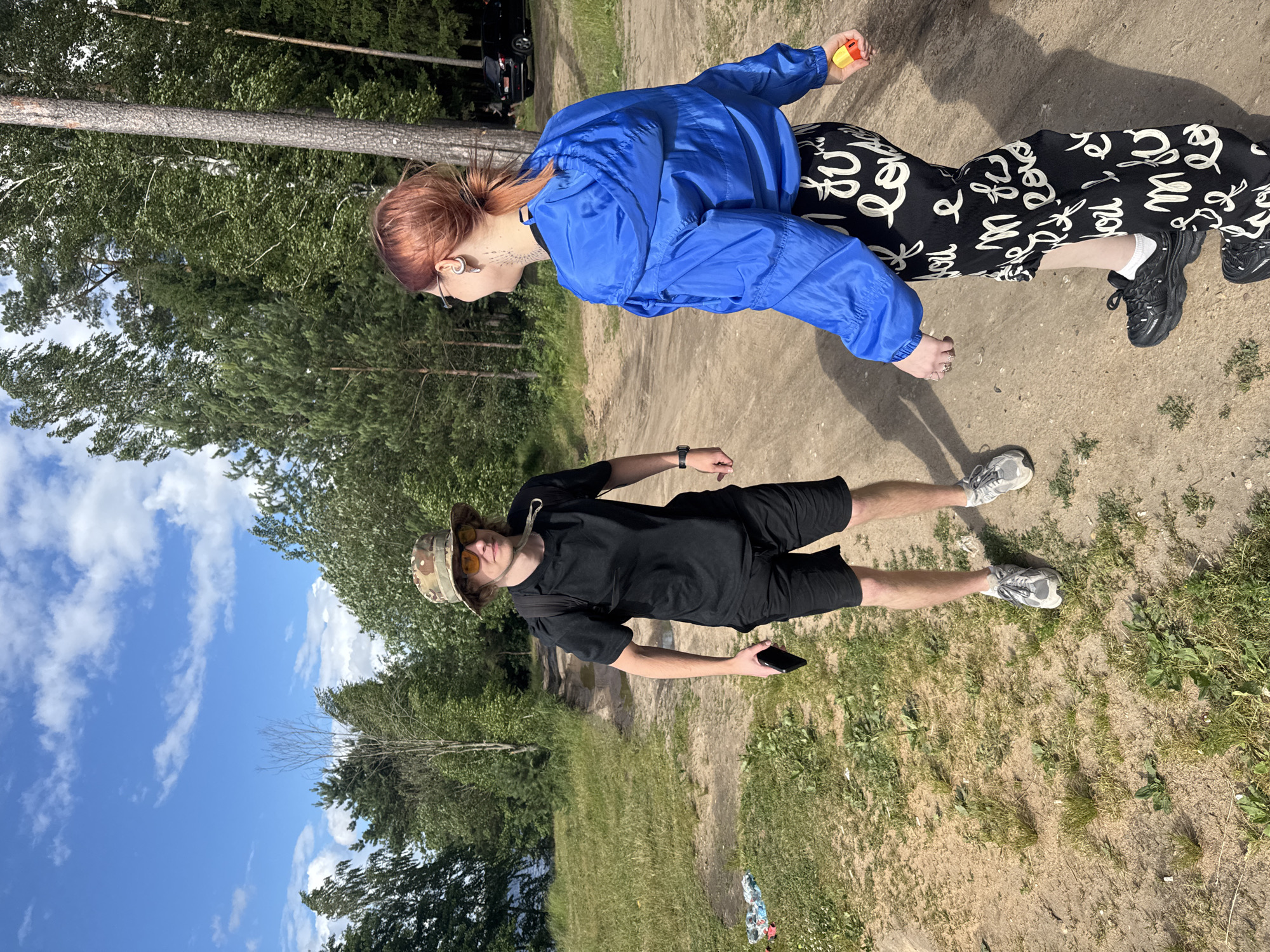 Two individuals are depicted outdoors on a dirt path bordered by green grass and trees. The image is oriented vertically.
In the foreground, a person wearing a black t-shirt, black shorts, a camouflage bucket hat, and sunglasses stands holding a smartphone. They are wearing athletic shoes.
In the background, another person is seen wearing a bright blue jacket, black pants with a white graphic print, and black sneakers. This person is holding a small yellow and red object. They are wearing earrings and glasses.
The scene appears to be a park or natural area with a dirt path, grass, and a dense treeline. The sky is blue with scattered white clouds. A black car is partially visible in the background near the trees. The lighting suggests it is daytime, likely sunny given the shadows. The overall atmosphere appears casual and relaxed. The provided location context is Desnogorsk, Russia.