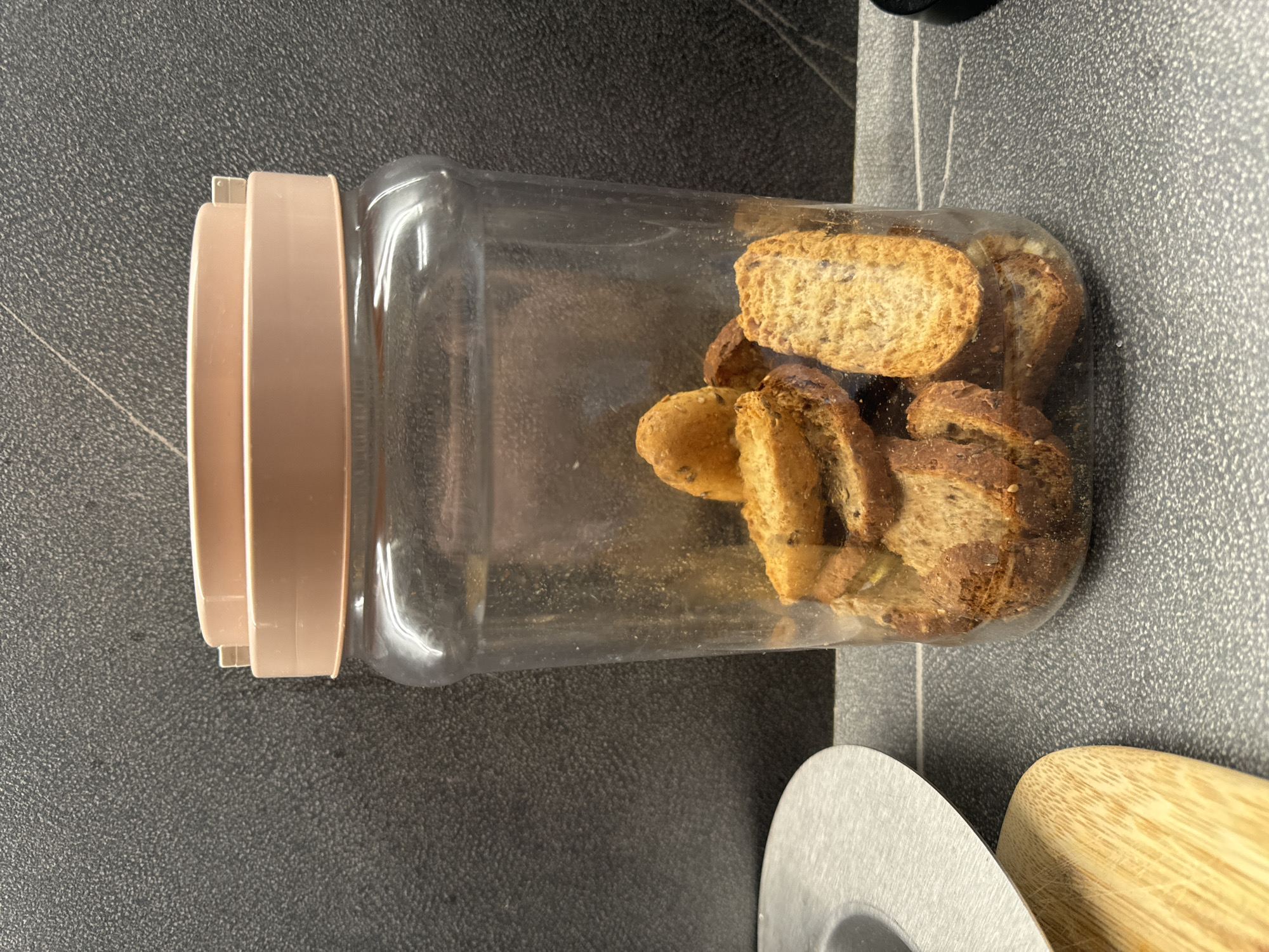 A clear plastic jar filled with toasted bread slices, possibly biscotti, sits on a speckled gray countertop. The jar has a light pink lid. To the left of the jar, a portion of a wooden cutting board and a metallic object, possibly a sieve or colander, are visible. The background is a dark gray textured wall. The scene appears to be in a kitchen environment, with no people or animals present. There are no specific landmarks or clues to indicate the location within Murcia, Spain.