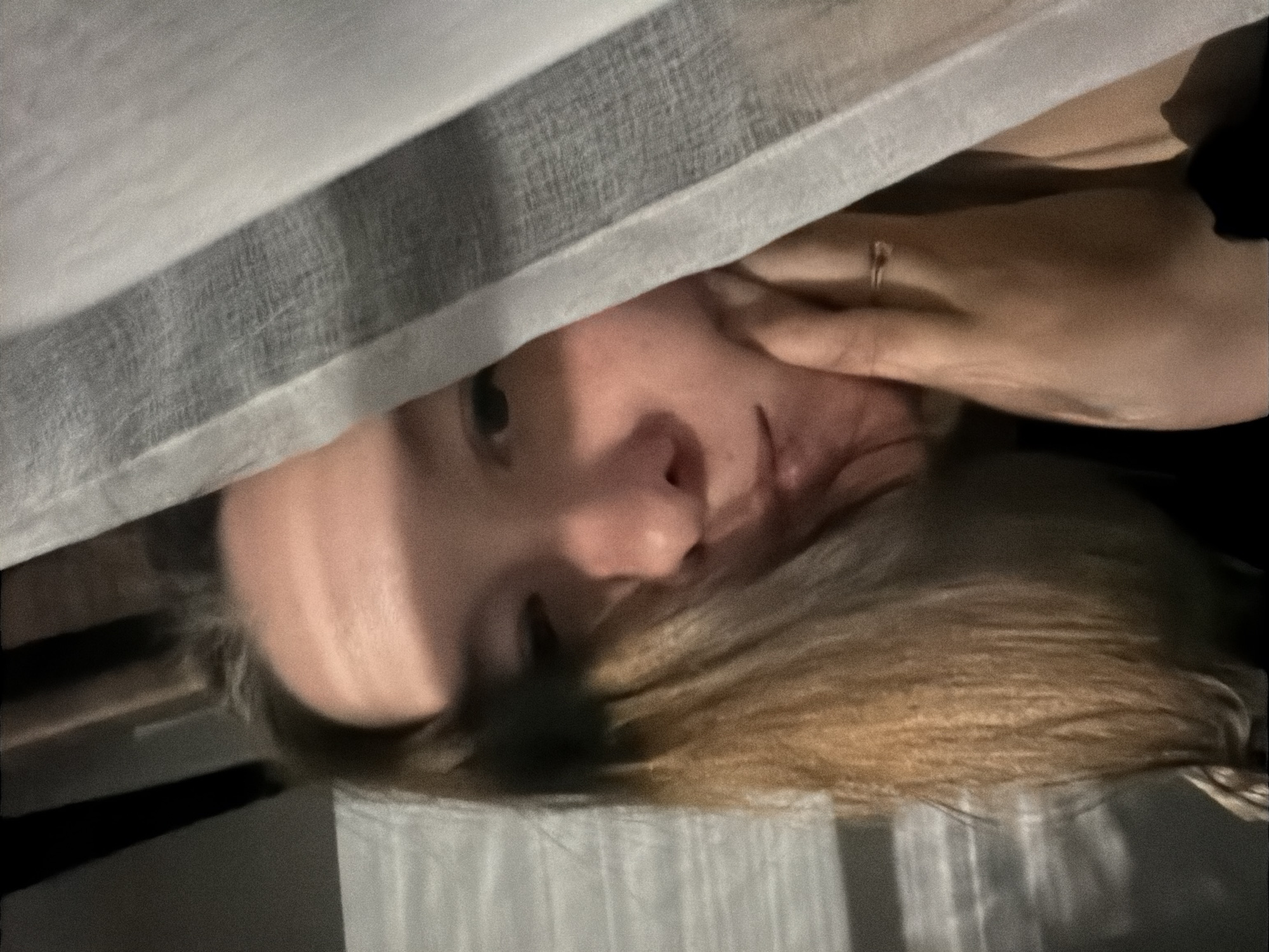 A person with blond hair is partially visible from under a grey curtain. The person's face is partially obscured by the curtain. A hand with a ring on the ring finger is touching the person's face. The background is dark. The image does not contain any specific landmarks indicating the city of Staraya, Russia.