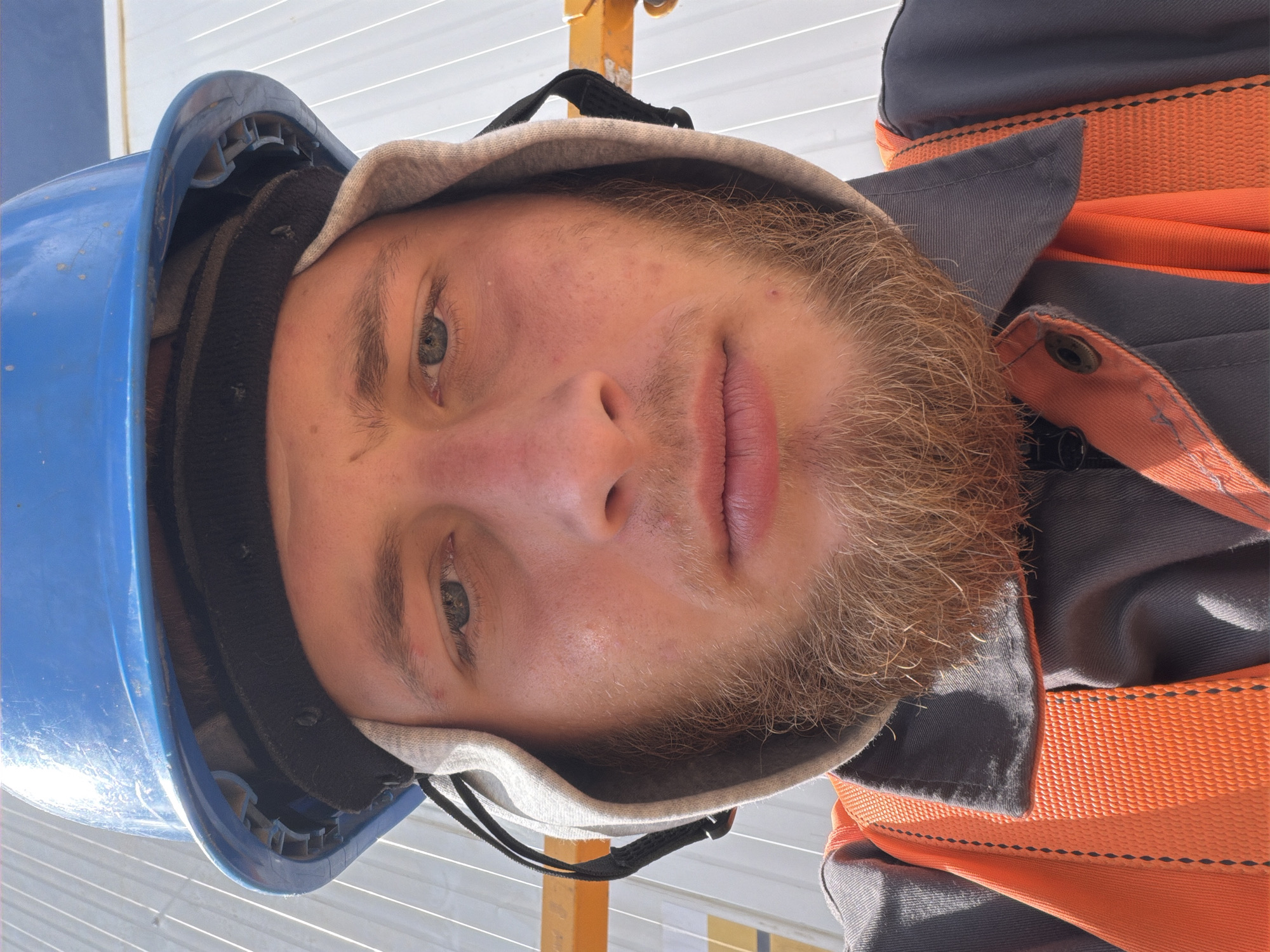 A young man with light brown hair and a beard, wearing a blue hard hat, grey work shirt, and an orange safety vest, is pictured. He is looking directly at the camera, with his face filling most of the frame. The background appears to be a construction site, with white siding and yellow scaffolding visible. The image is a selfie, taken from a low angle. There is no indication of the location being Wilkowisko, Poland.