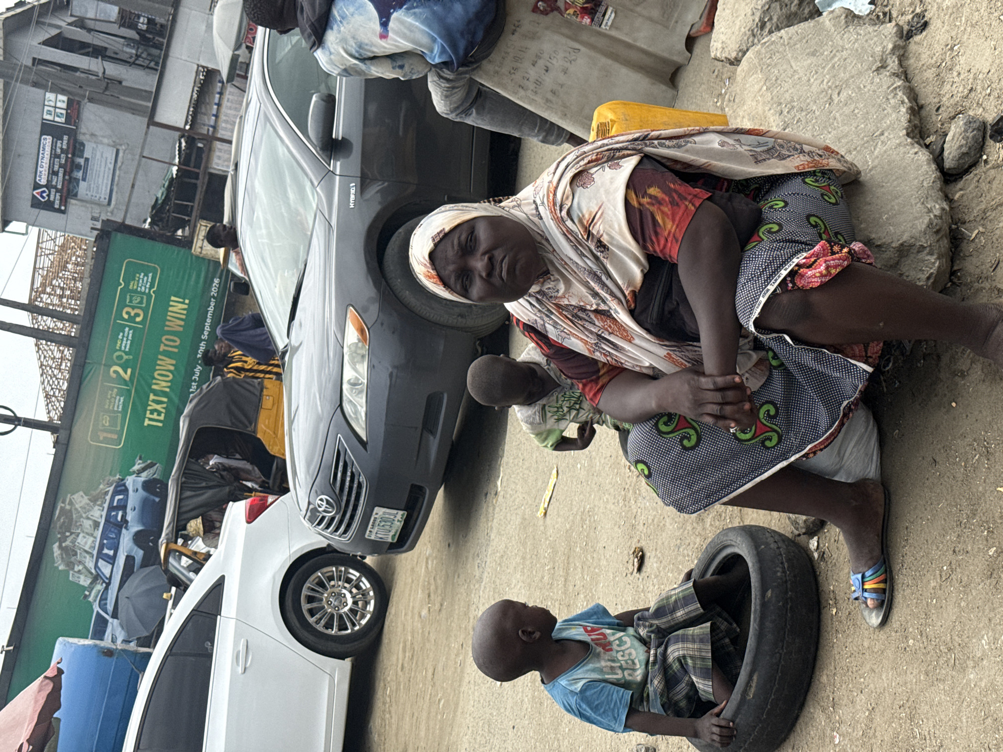 In Lekki, Nigeria, a woman in a headscarf and patterned clothing sits on a large rock. Beside her, a child in a blue t-shirt and checkered shorts is seated inside a tire. Another child in a green and white shirt is visible behind them. In the background, cars are present, including a dark grey sedan and a white car, with a yellow motorized tricycle partially visible. A large green billboard with text and images of vehicles and money is also prominent in the background. The scene appears to be on a dirt road.
