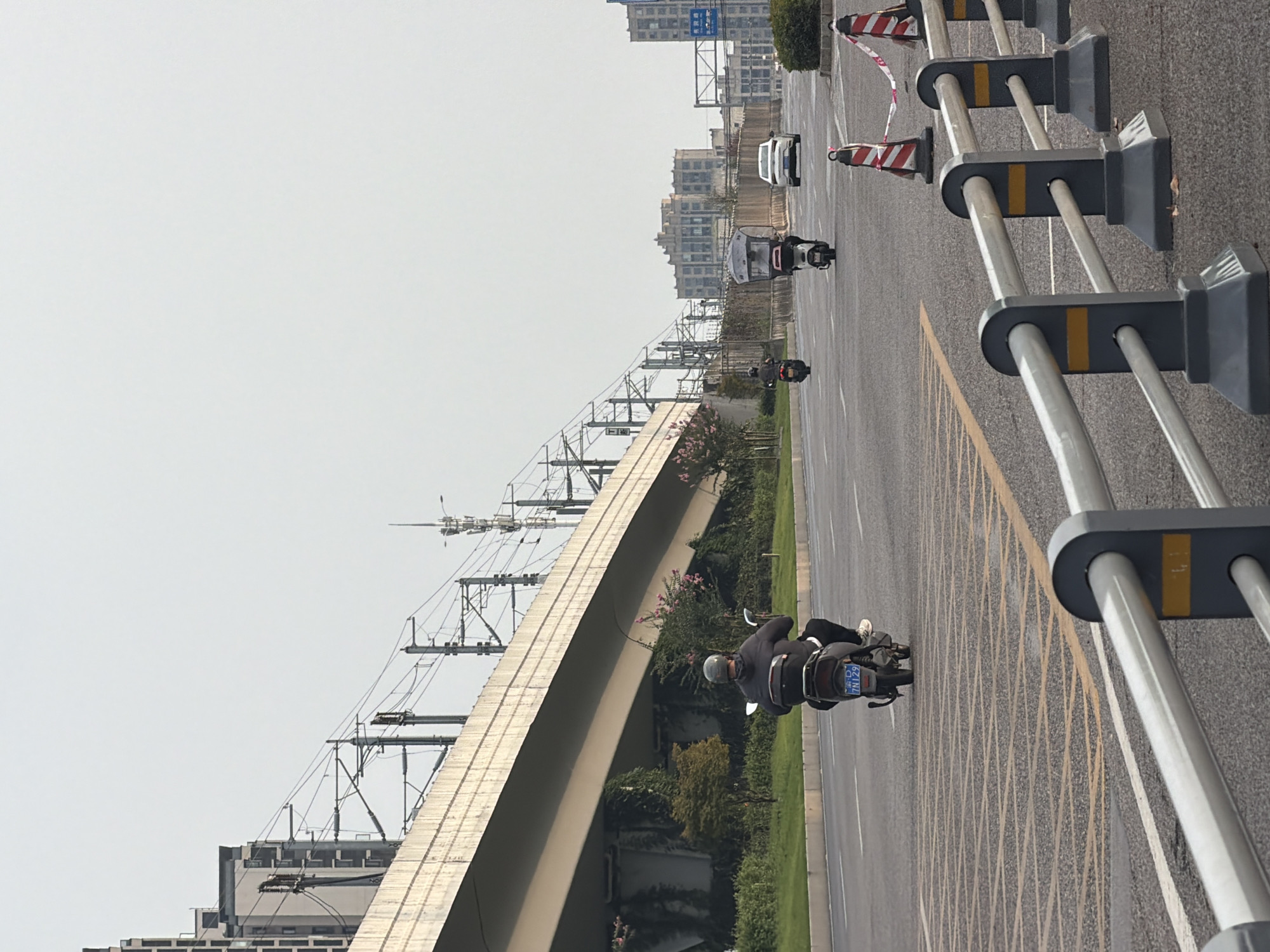 The image depicts a road scene in Jiangjin, China, characterized by an elevated overpass with train tracks and overhead electrical lines. A person is riding a scooter on the right side of the road, facing away from the camera. In the distance, two other scooters and a white car are visible. The road is marked with yellow lines and features a safety barrier in the foreground. Trees and buildings are present in the background.