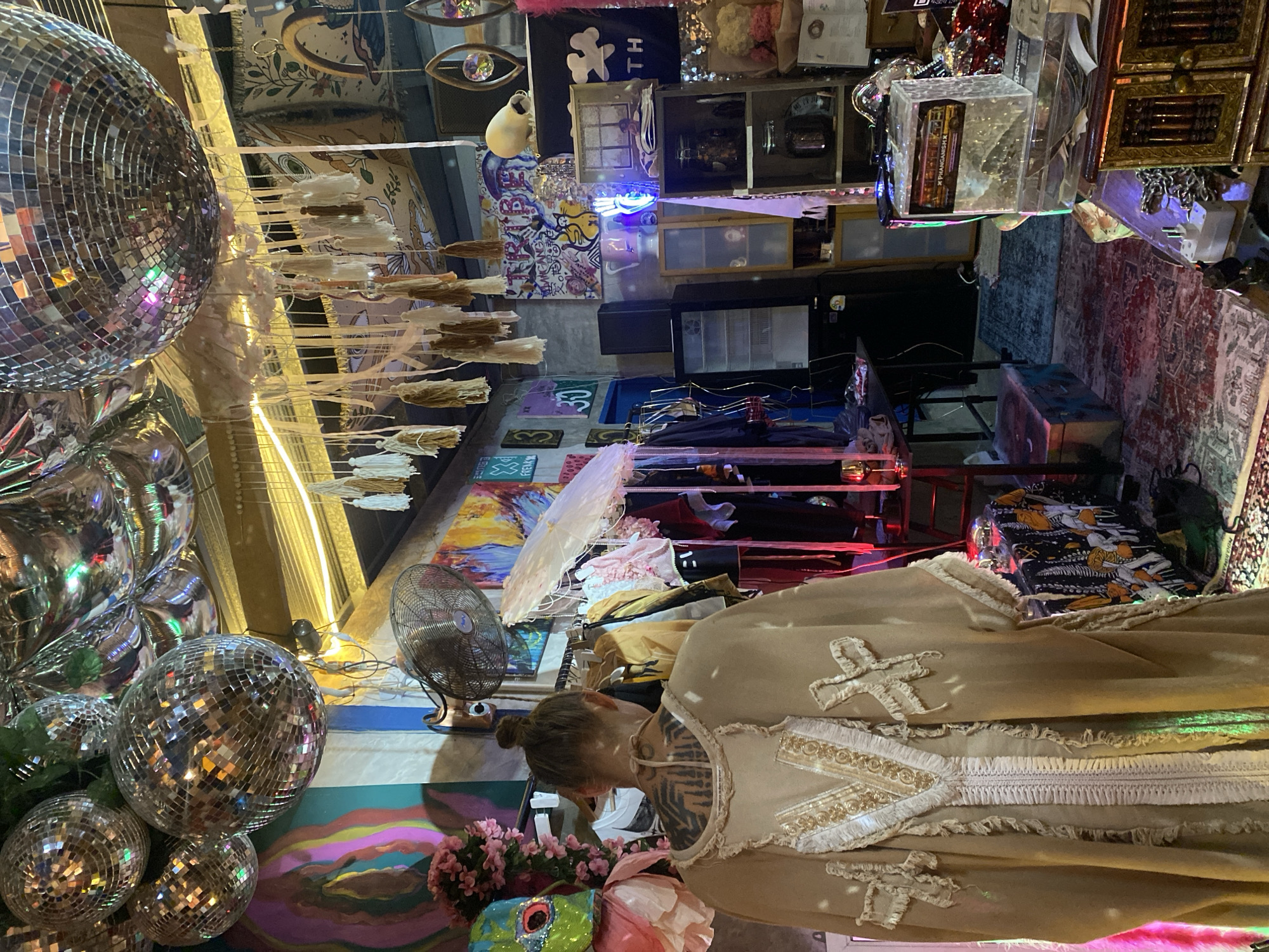 A person wearing a beige dress with ornate trim is standing in a cluttered indoor space. The location appears to be in Baan Nai Suan, Thailand, given the context. The background is filled with assorted objects including disco balls, fans, artwork, shelves, and lighting. The lighting is a mix of natural and artificial. The person is partially obscured by the dress.